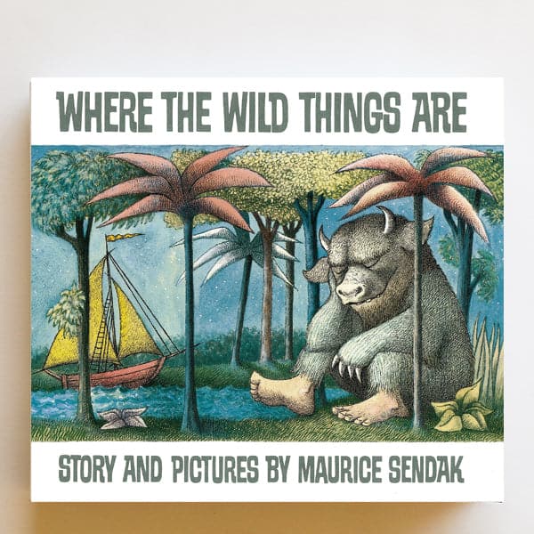 This classic children's story book features a dreamy forest homing a large mystic creature with pale blue fur, sharpened claws and round horns. The creature sits peacefully among the trees and across from a pond filled with a soft yellow and red sail boat. The book is titled 'Where the Wild Things Are' in pale teal capital lettering above. 
