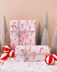 A single sheet of gift wrap with a blush background and a repeating red and white candy cane print. The gift wrap is reversible and the other side features a light purple / lavender background with an all over red and white candy cane design but the candy canes are slightly smaller than the reverse. Present is wrapped with a silver tinsel ribbon and surrounded by snow, red and white ornaments and silver tinsel trees.