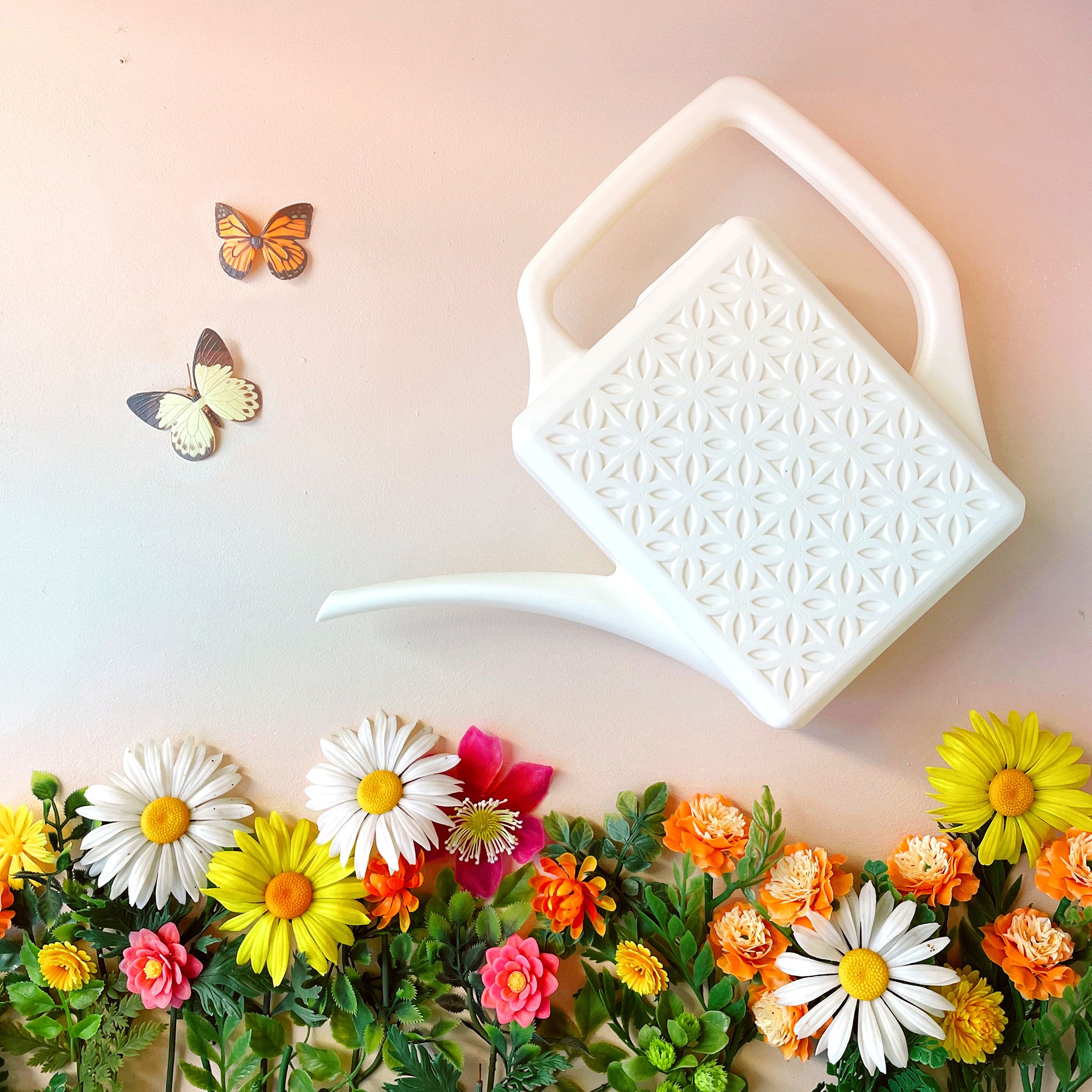 An ivory plastic watering can with a narrow spout and square handle and a rectangle breeze block design on the sides. This photograph has the watering can staged above a bed of pink, yellow and white florals and two butterflies on the left hand side.