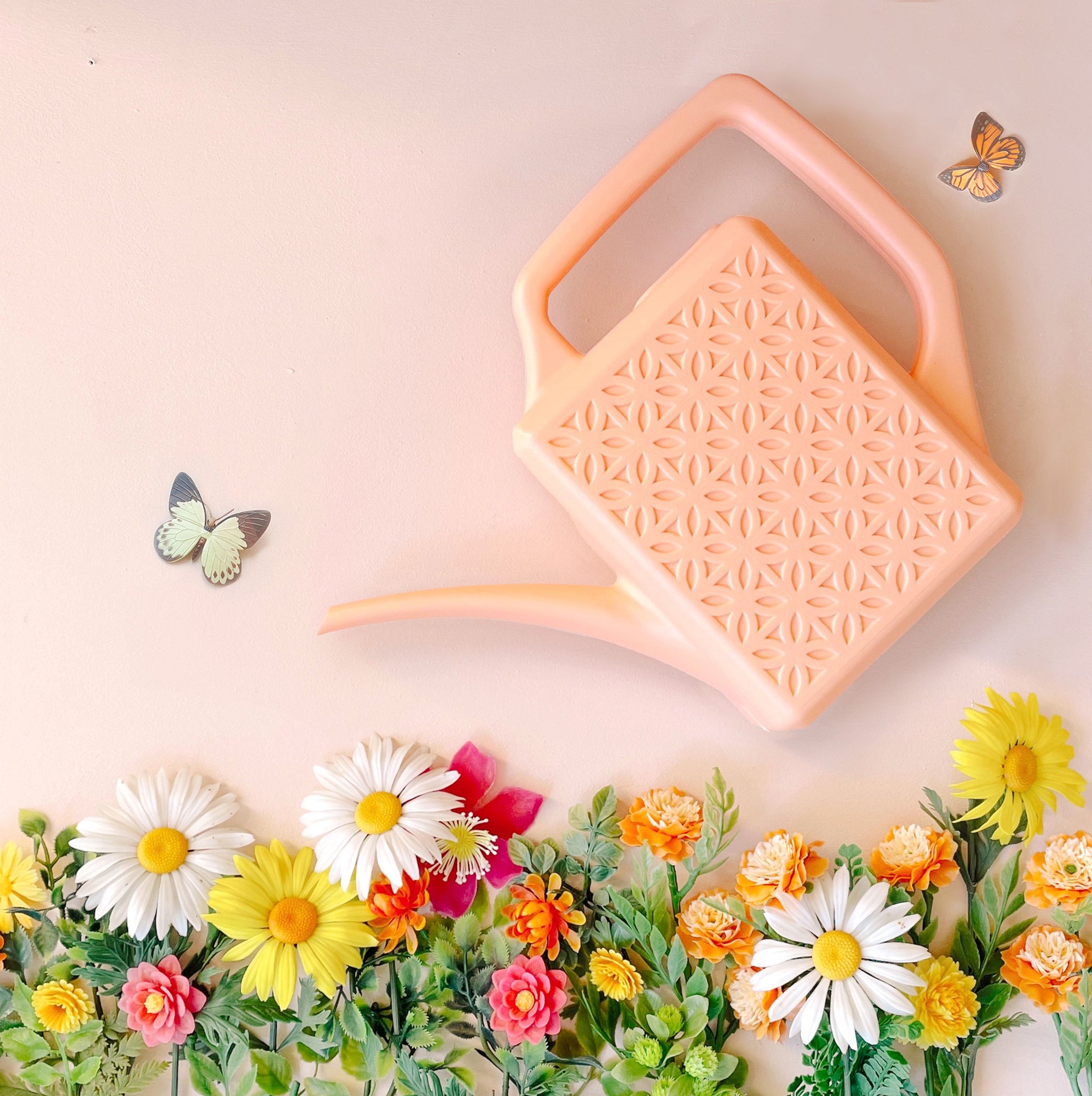 A peach plastic watering can with a narrow spout and square handle and a rectangle breeze block design on the sides. This photograph has the watering can staged above a bed of pink, yellow and white florals and two butterflies on each side of the can.