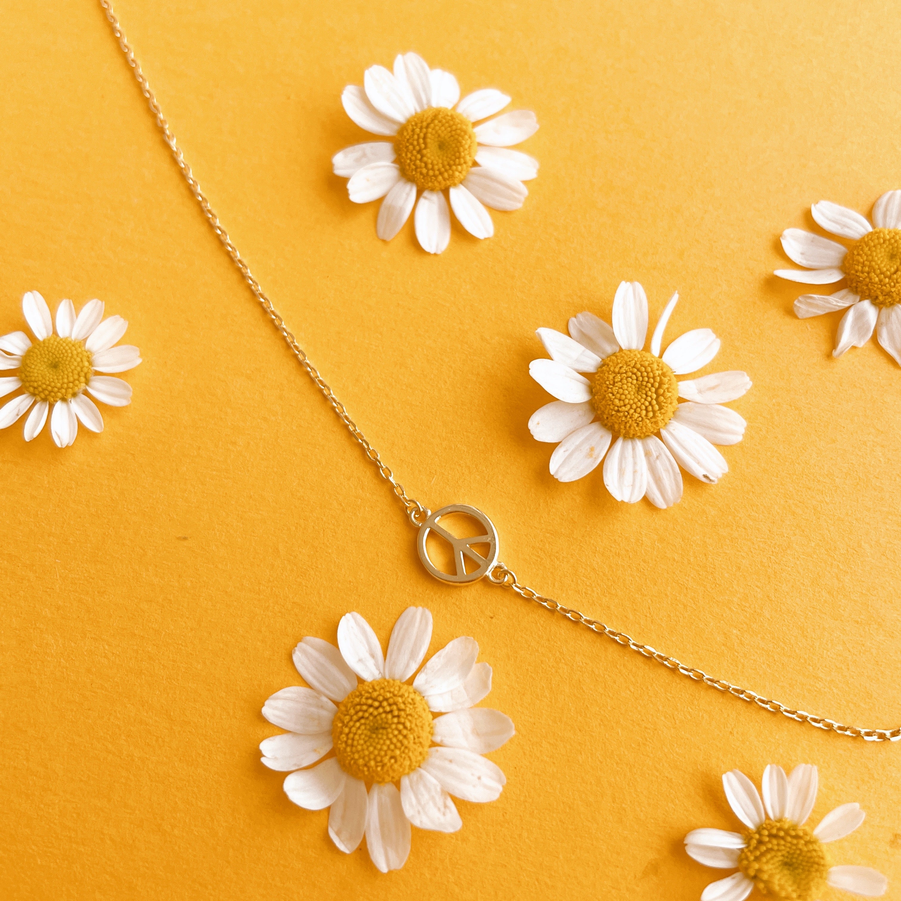 Gold chain necklace complete with classic peace sign symbol pendant. The pendent is connected to the chain by two loops on each side.   In this image the necklace is draped on a bright yellow paper with daisies sprinkled around it like confetti.