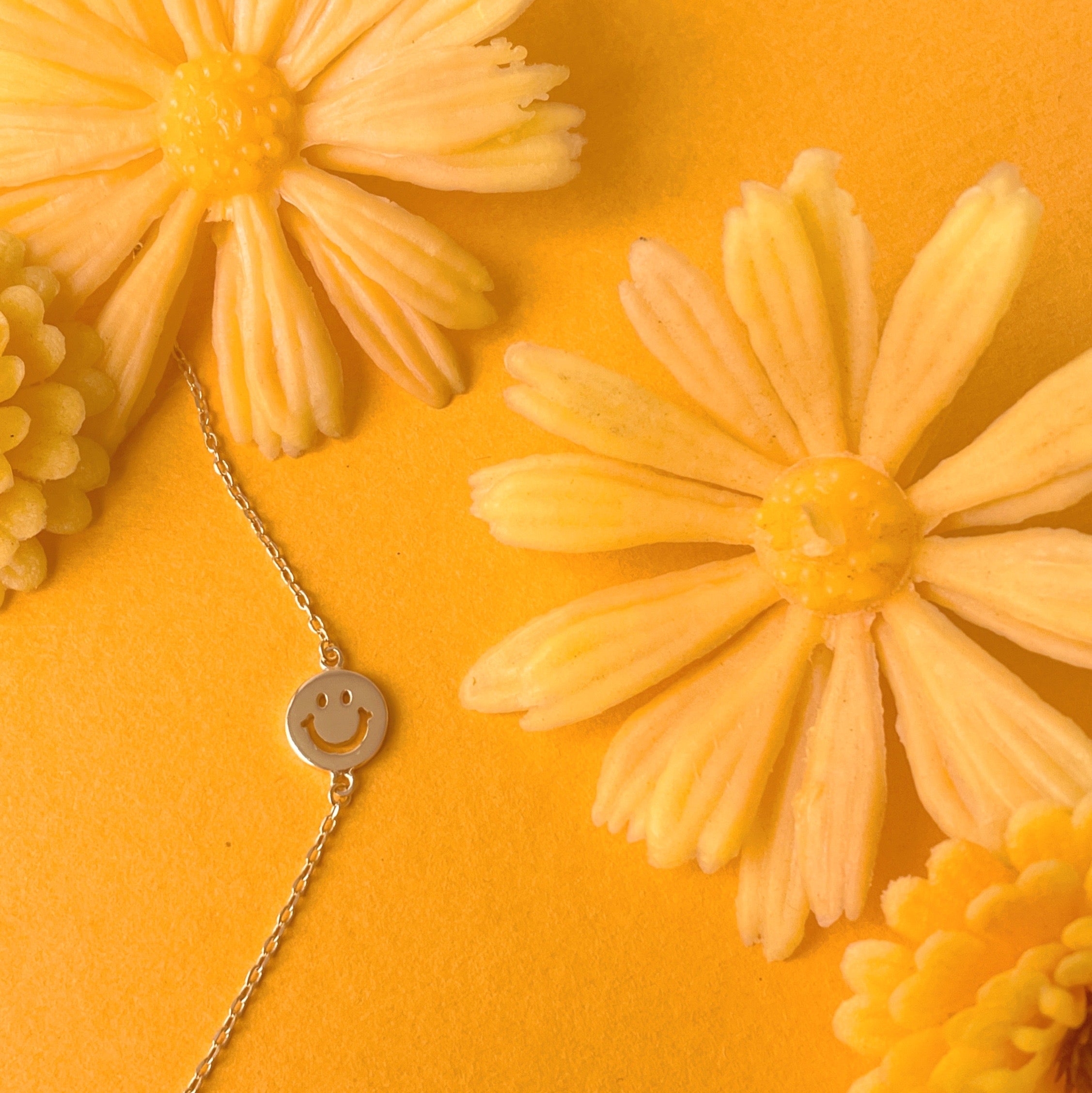 Gold chain necklace complete with classic smiley face pendant turned sideways. The pendant is connected to the chain by two loops on each side.  This image features the necklace resting on a marigold colored ground with fake yellow daisies surrounding it.