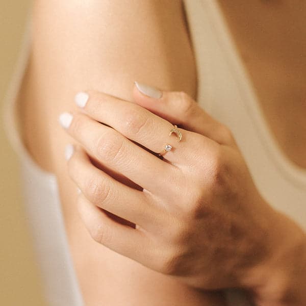 A gold ring with an open front that features a cubic zirconia on one end and a crescent moon on the other, photographed worn on a models hand. 
