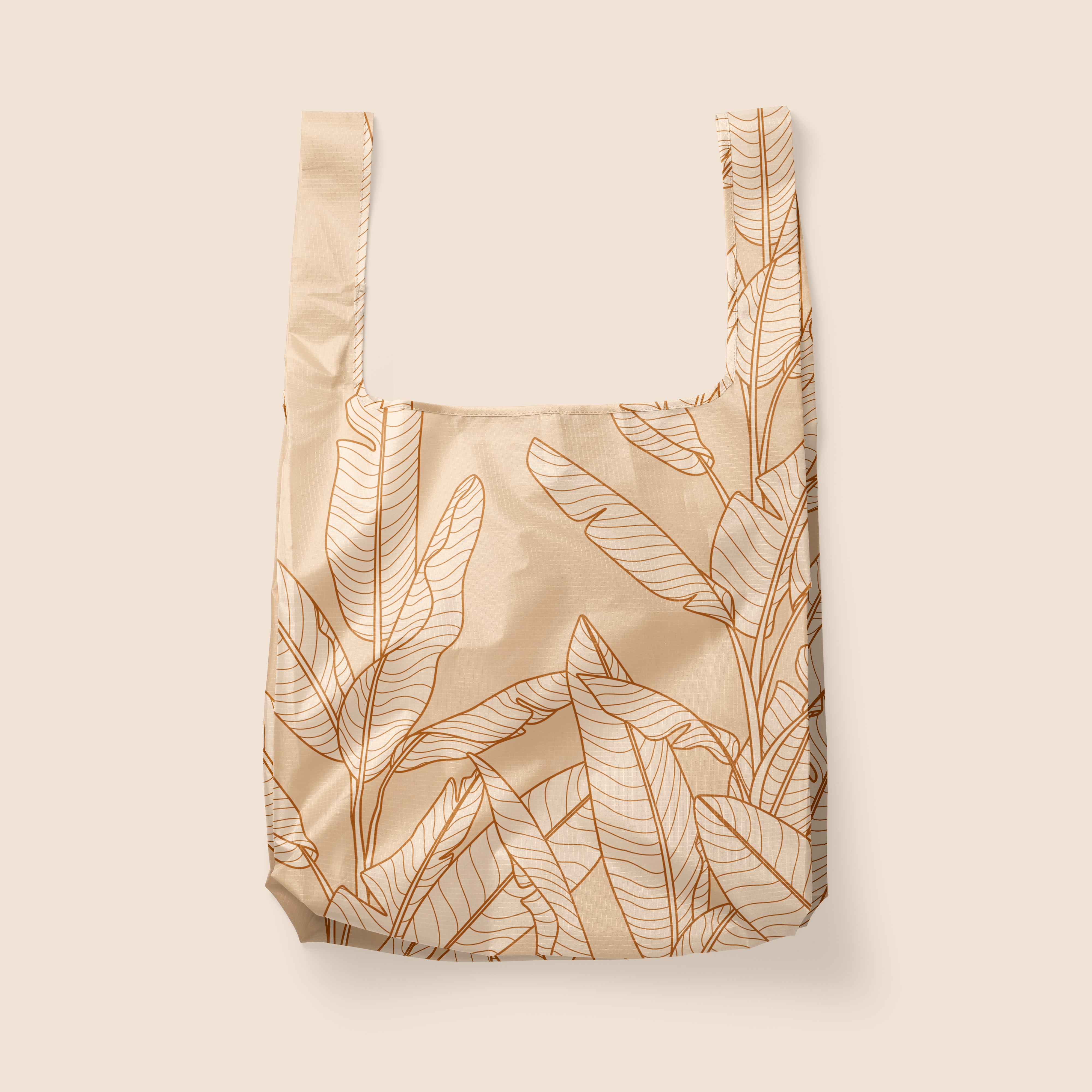 Photographed in front of a white background is a light tan / pink nylon reusable bag with a cream outlined Banana Leaves print on the front and back.