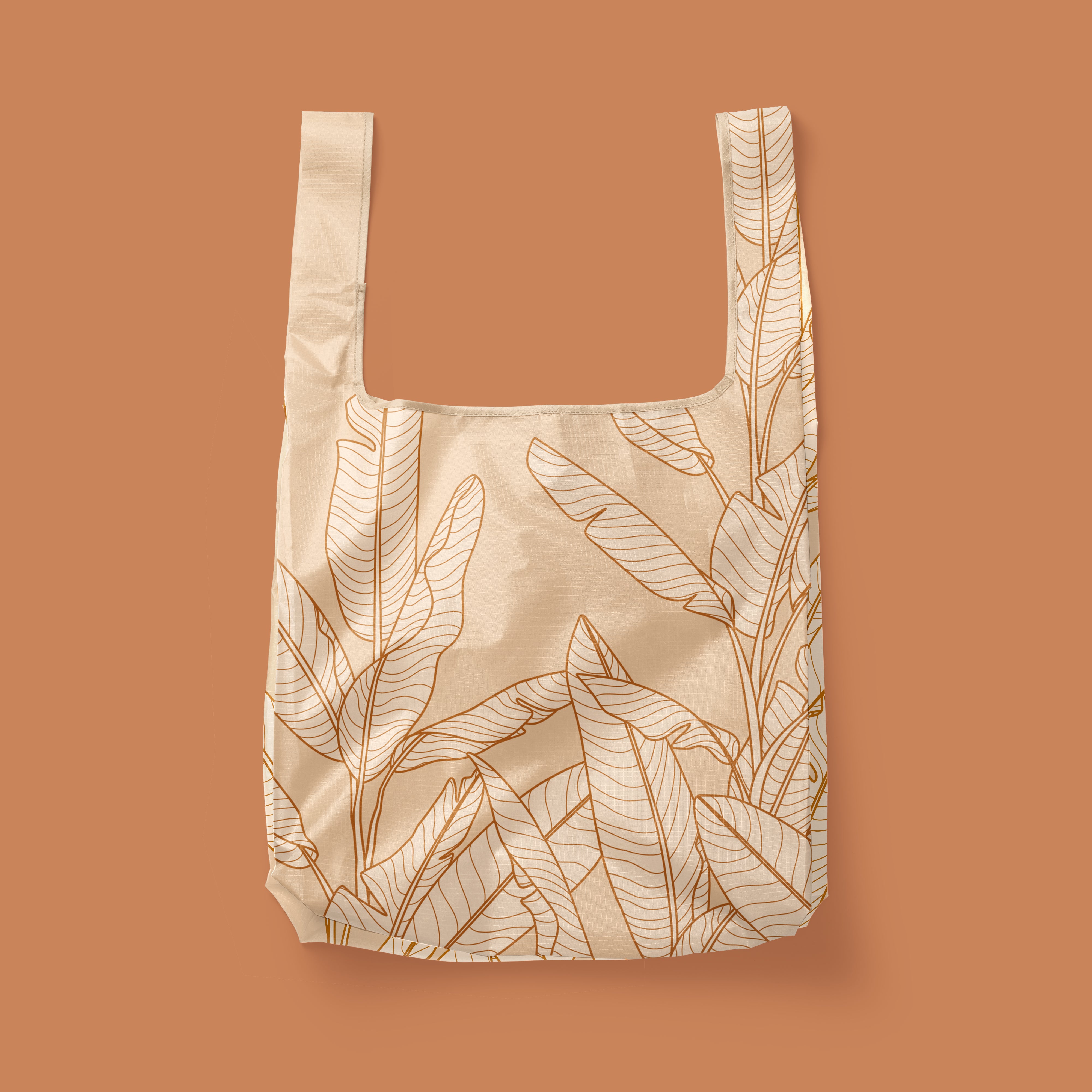 Photographed in front of a terracotta background is a light tan / pink nylon reusable bag with a cream outlined Banana Leaves print on the front and back.