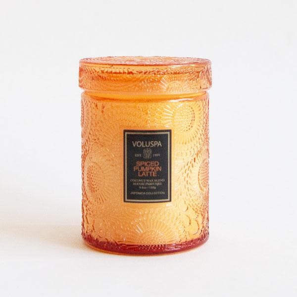 In front of a white background is an orange glass, cylinder jar. The glass has a floral texture. On the front is a black sticker with gold text that reads ‘voluspa.’ Below is orange text that reads ‘spiced pumpkin latte.’ On top of the jar is a matching glass lid. 