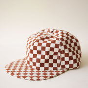 A white and rust checkered six panel hat and features a fabric strap with brass metal clasp.