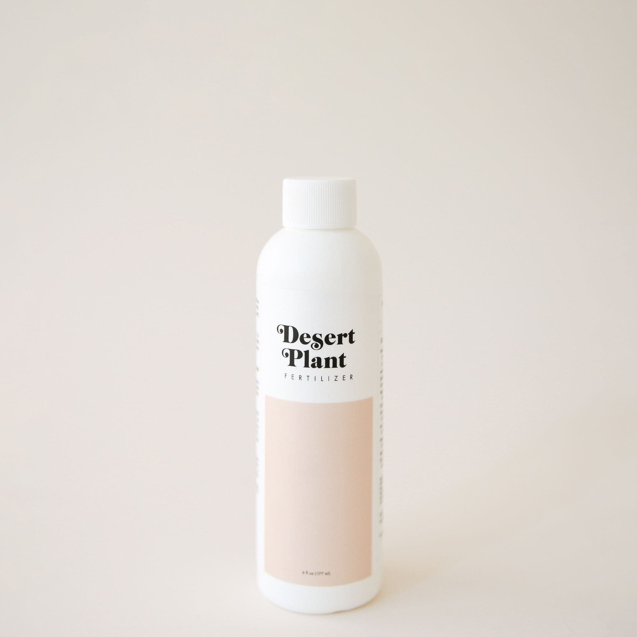 White twist top bottle labeled &#39;Desert Plant Fertilizer&#39; in black playful lettering. Below the label is a rectangular block of soft beige. The fertilizer sits in front of a solid sand colored background.