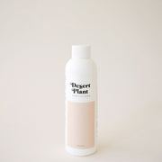 White twist top bottle labeled 'Desert Plant Fertilizer' in black playful lettering. Below the label is a rectangular block of soft beige. The fertilizer sits in front of a solid sand colored background.