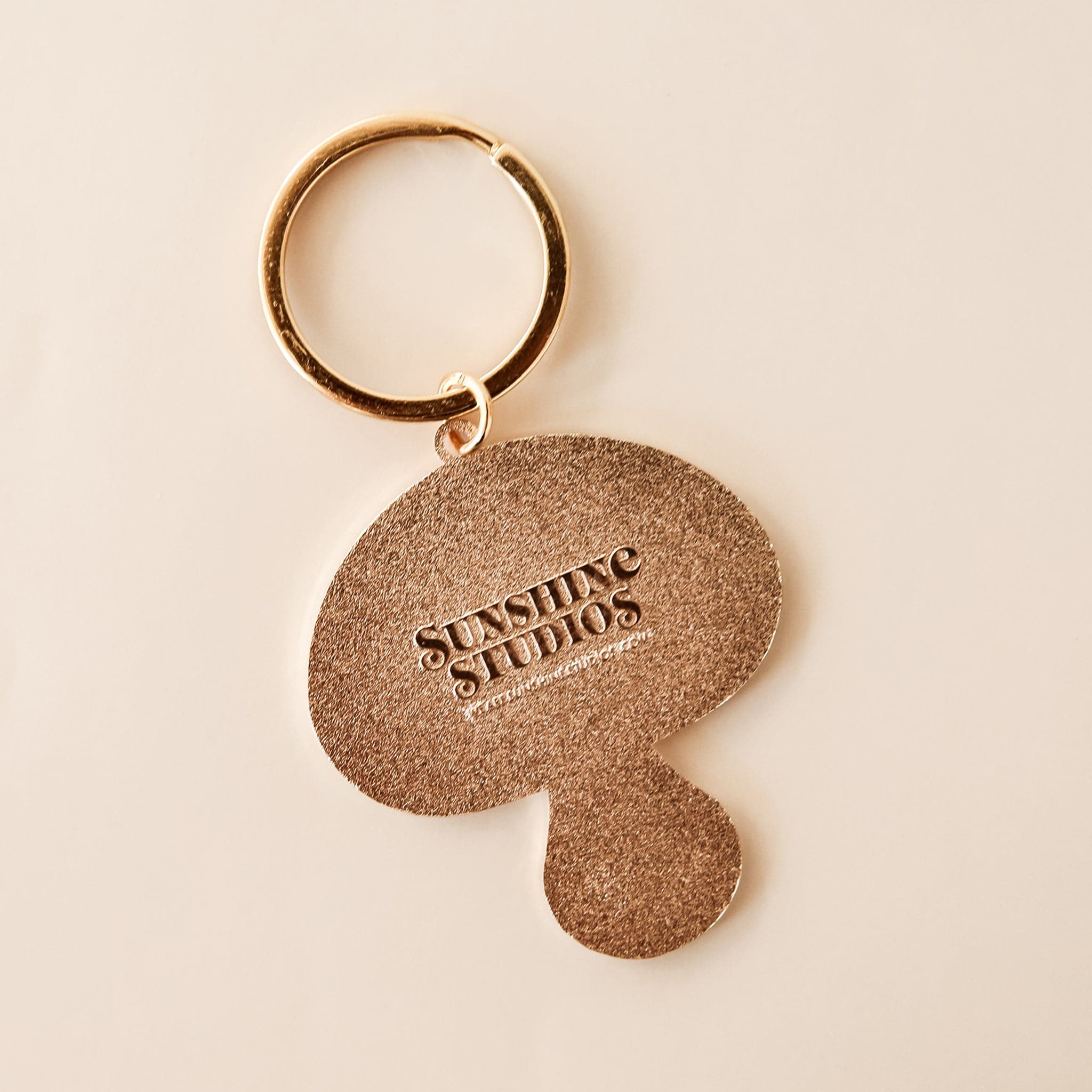 Backside of mushroom keychain is golden and textured, reading &#39;sunshine studios&#39; in raised lettering. 