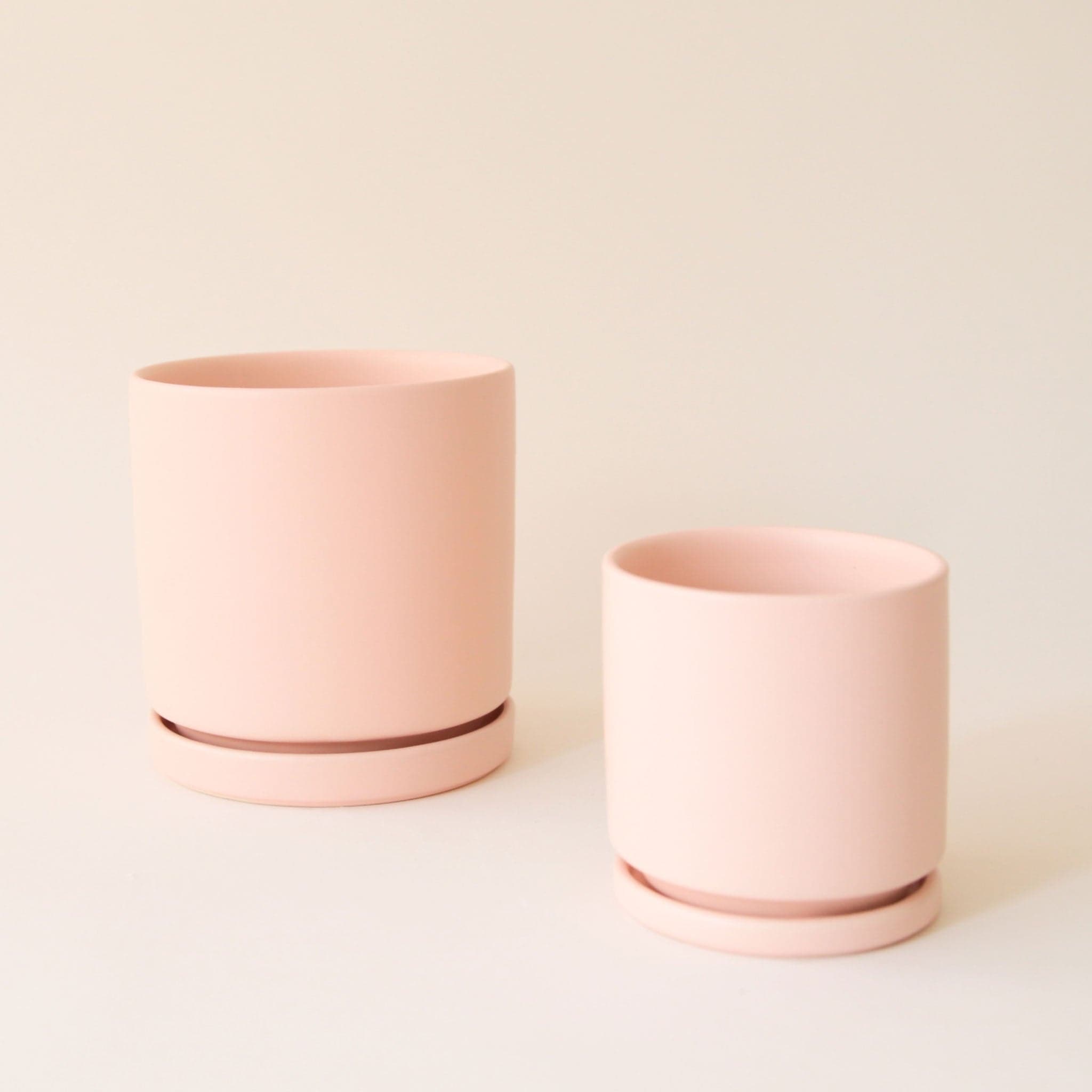 On a cream background is two different sized ceramic pots in a light pink shaded with removable trays for watering.