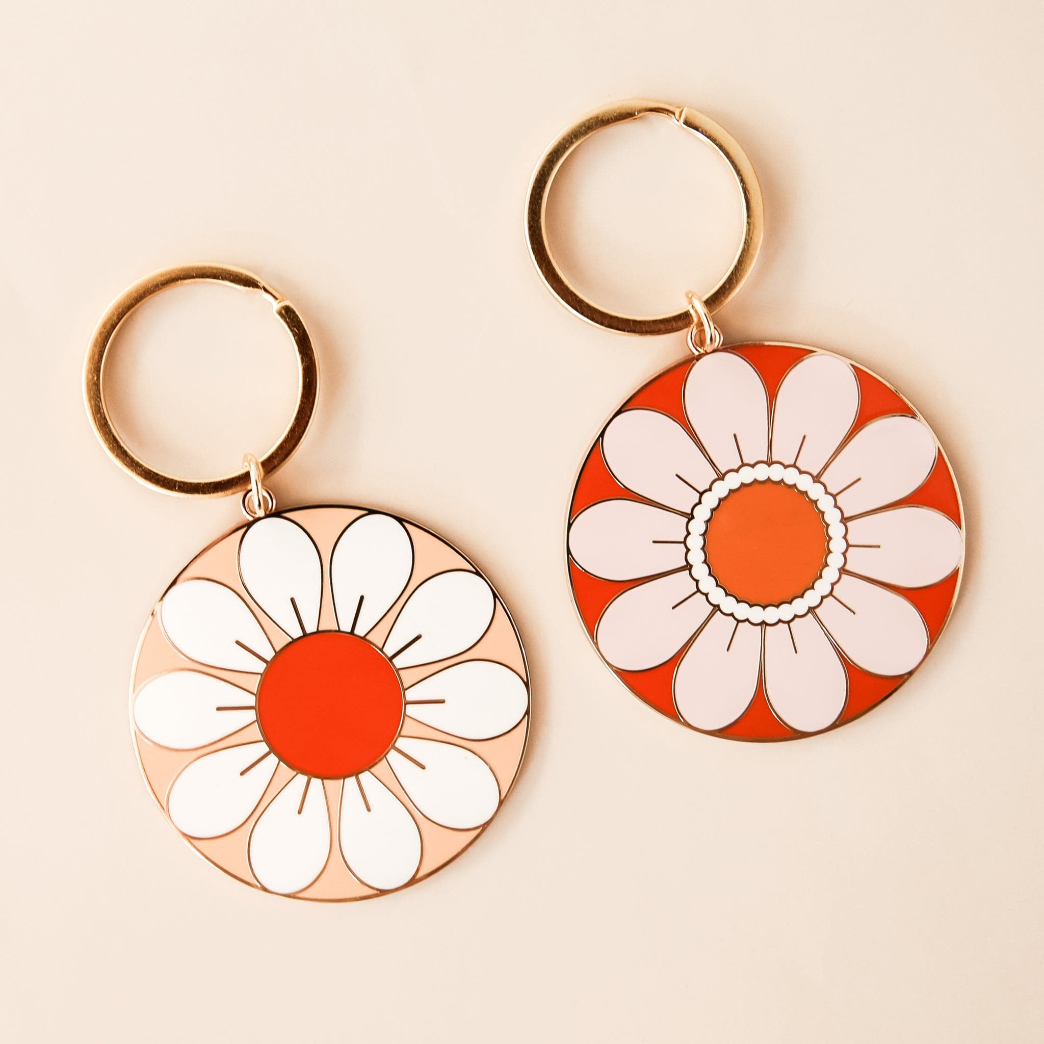 Two circular flower key chains both complete with golden key chain rings. To the left is a flower with white petals and red center and to the right is a flower with light beige petals and orange center. 