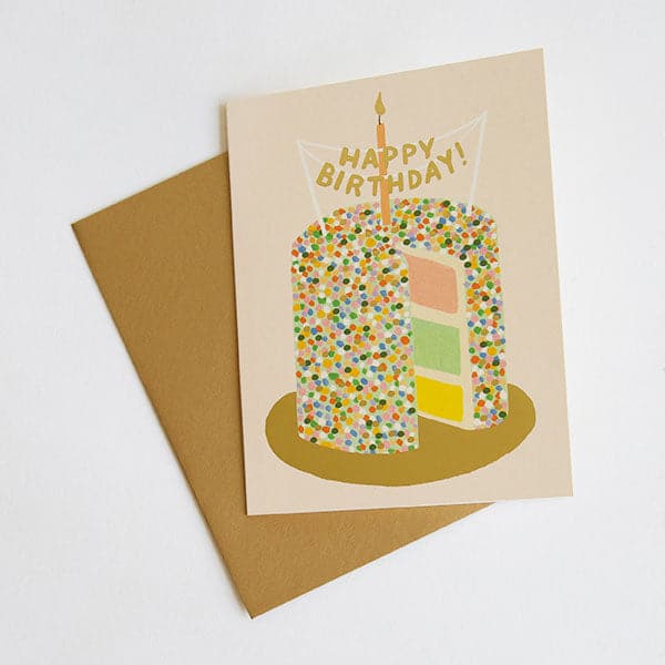 On a white background is a light pink card with a multicolored cake illustration on the front that has a single candle and a happy birthday sign on top. Also included is a coordinating gold envelope.