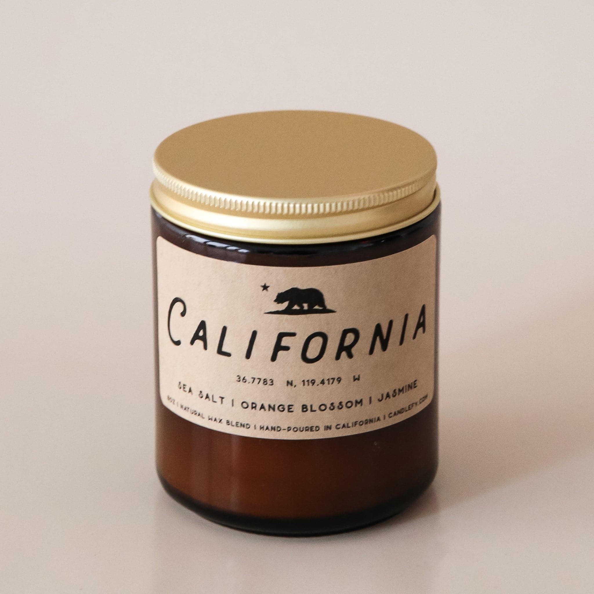An amber glass jar candle with a light tan rectangular label that reads, "California Sea Salt | Orange Blossom | Jasmine |" in black letters along with a black filled in image of the California state bear and a gold screw on lid.