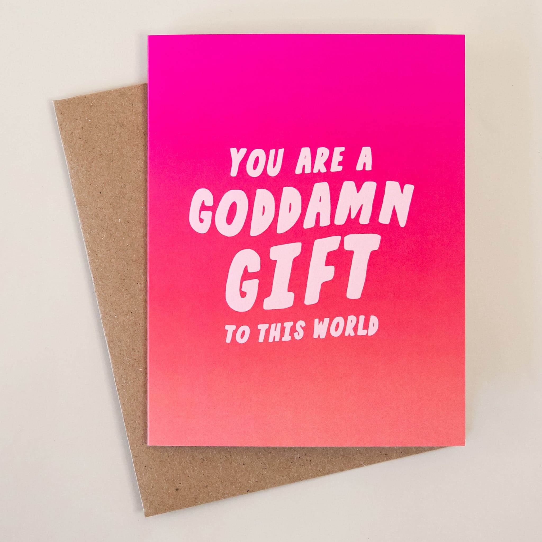 This card reads &#39;You Are a Goddamn Gift to This World&#39; with a background featuring an ombre of rosie tones. This card is accompanied by a kraft brown envelope.