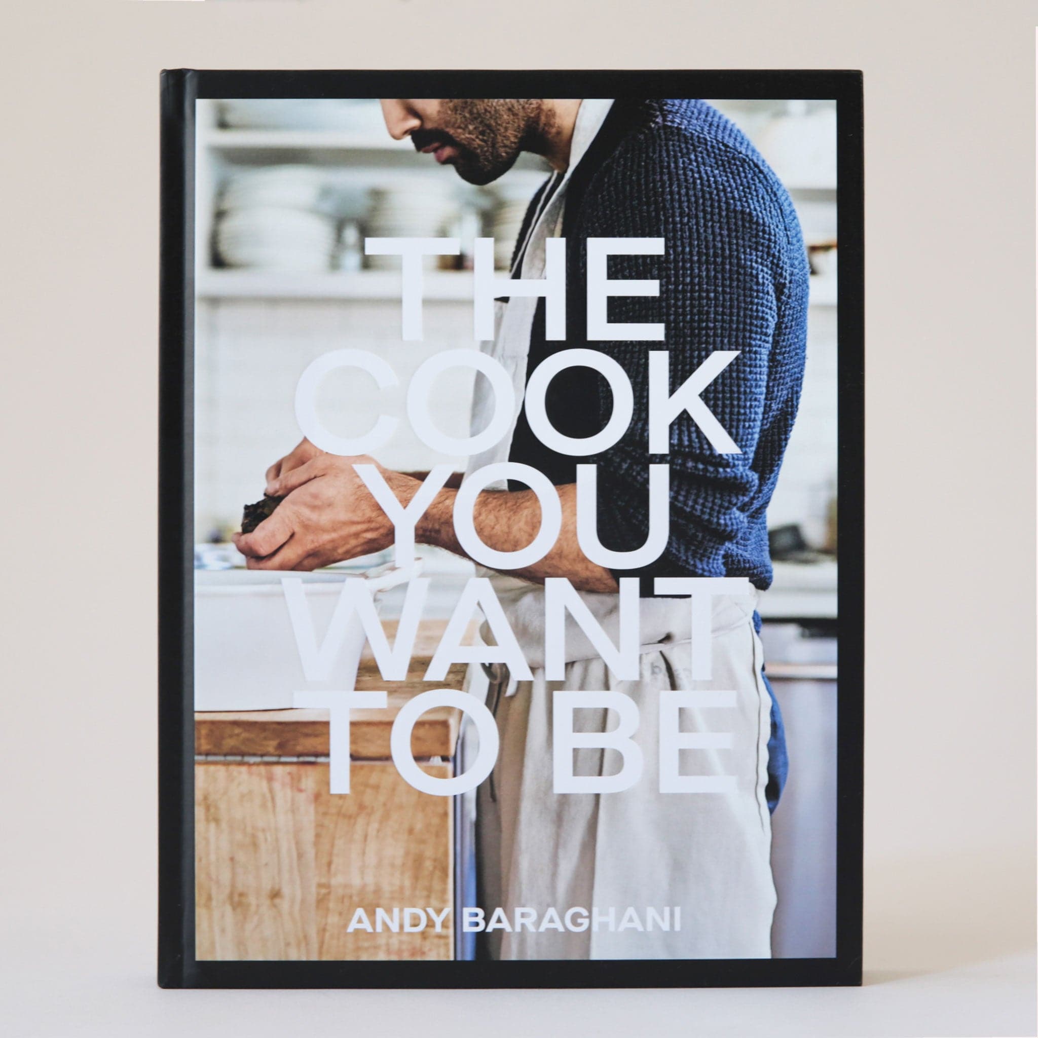 A sleek and stunning cookbook with the title, "The Cook You Want To Be" in white, bold text in front of a photo of a chef with an apron on, cooking.