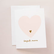 On a light pink background is a white card and envelope with a light pink heart in the center and a smaller gold heart inside of that along with gold text that reads, "Congrats Mama" along the bottom.