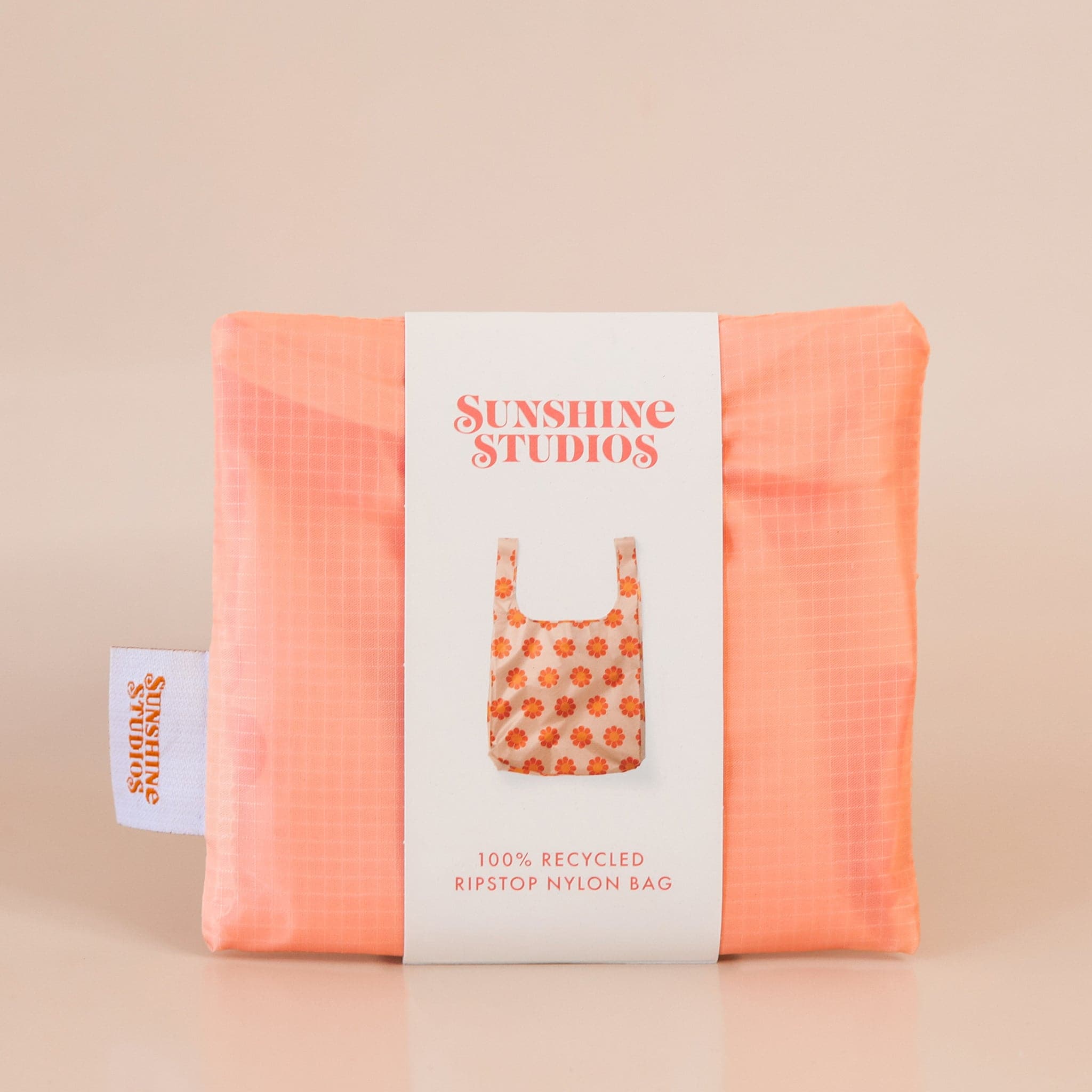 Peach reusable bag folded tightly into a square. The bag is wrapped in a white band that reads ’sunshine studios'. Under the text is a picture of the reusable bag. On the left side of the bag is a white tag with bright orange text that reads ’sunshine studios.' 