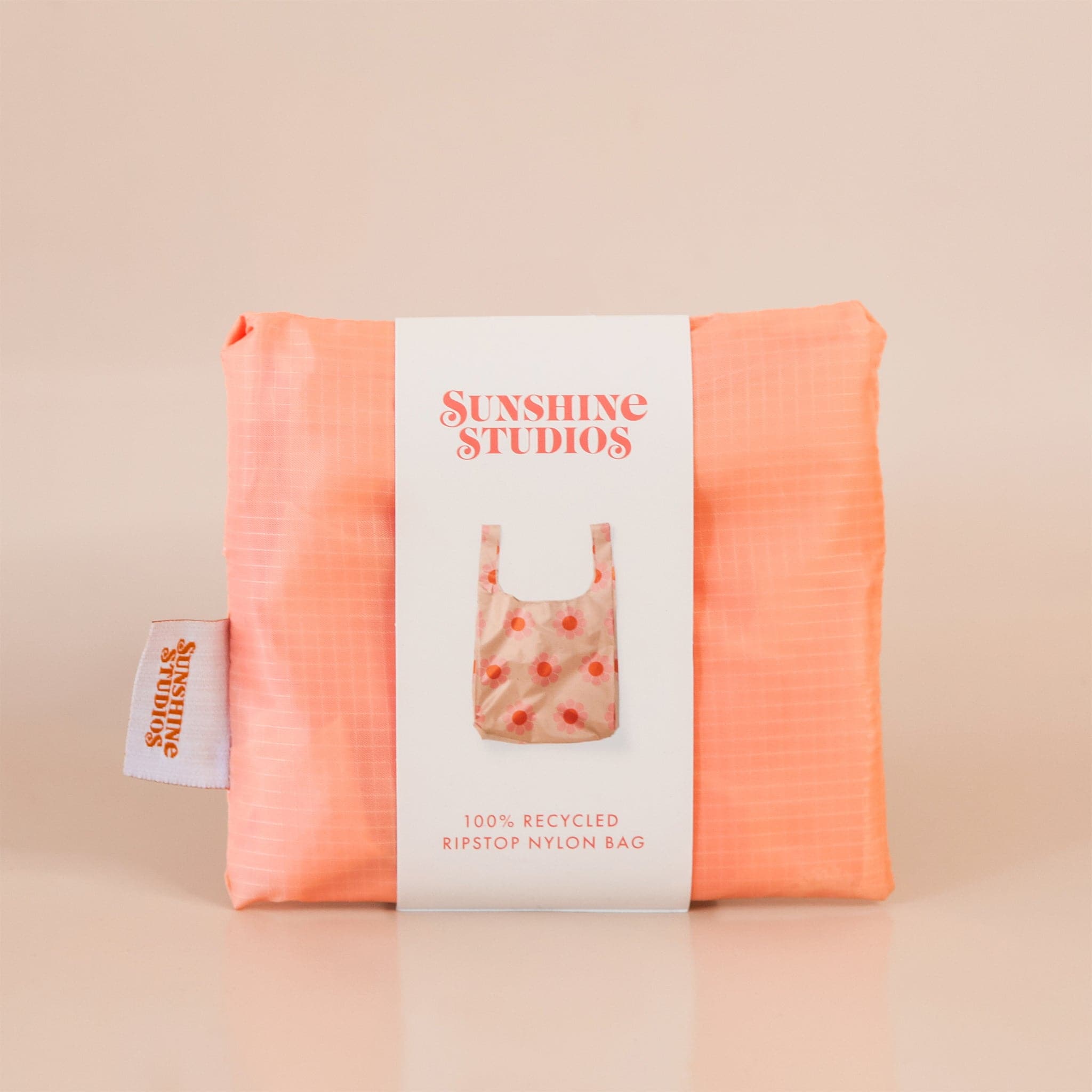 Peach reusable bag folded tightly into a square. The bag is wrapped in a white band that reads ’sunshine studios'. Under the text is a picture of the reusable bag. On the left side of the bag is a white tag with bright orange text that reads ’sunshine studios.'