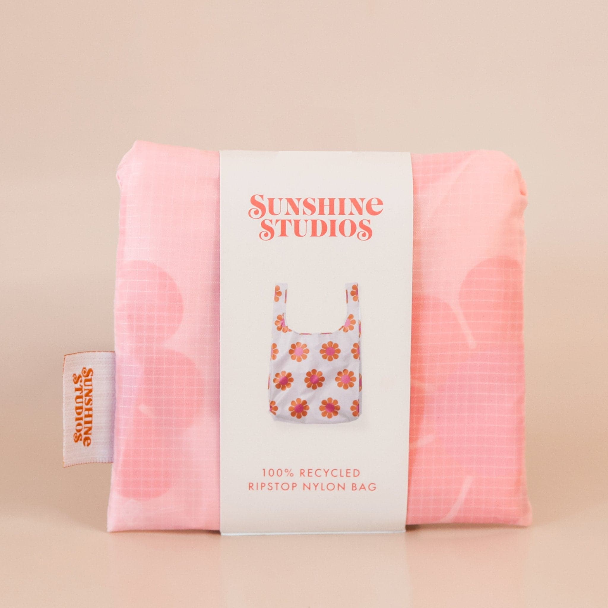 Soft pink reusable bag folded tightly into a square. The bag is wrapped in a white band that reads ’sunshine studios'. Under the text is a picture of the reusable bag. On the left side of the bag is a white tag with bright orange text that reads ’sunshine studios.'