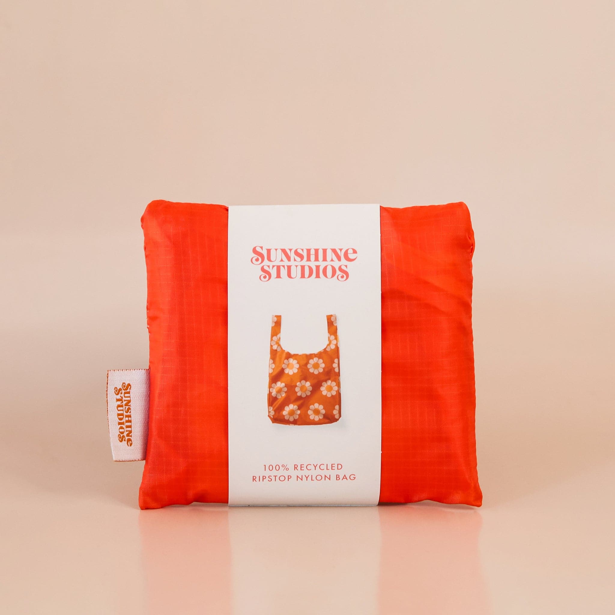 Folded up red-orange reusable bag folded into a small square within white wrap around packaging that reads &#39;sunshine studios&#39;. The label has a photo of the bag unfolded and reads &#39;100% recycled ripstop nylon bag&#39;