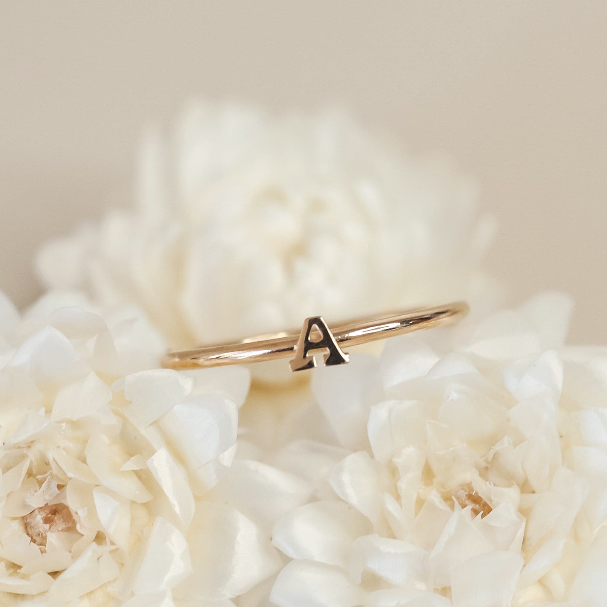 On a cream background is a dainty gold ring with an &quot;A&quot; in the center.