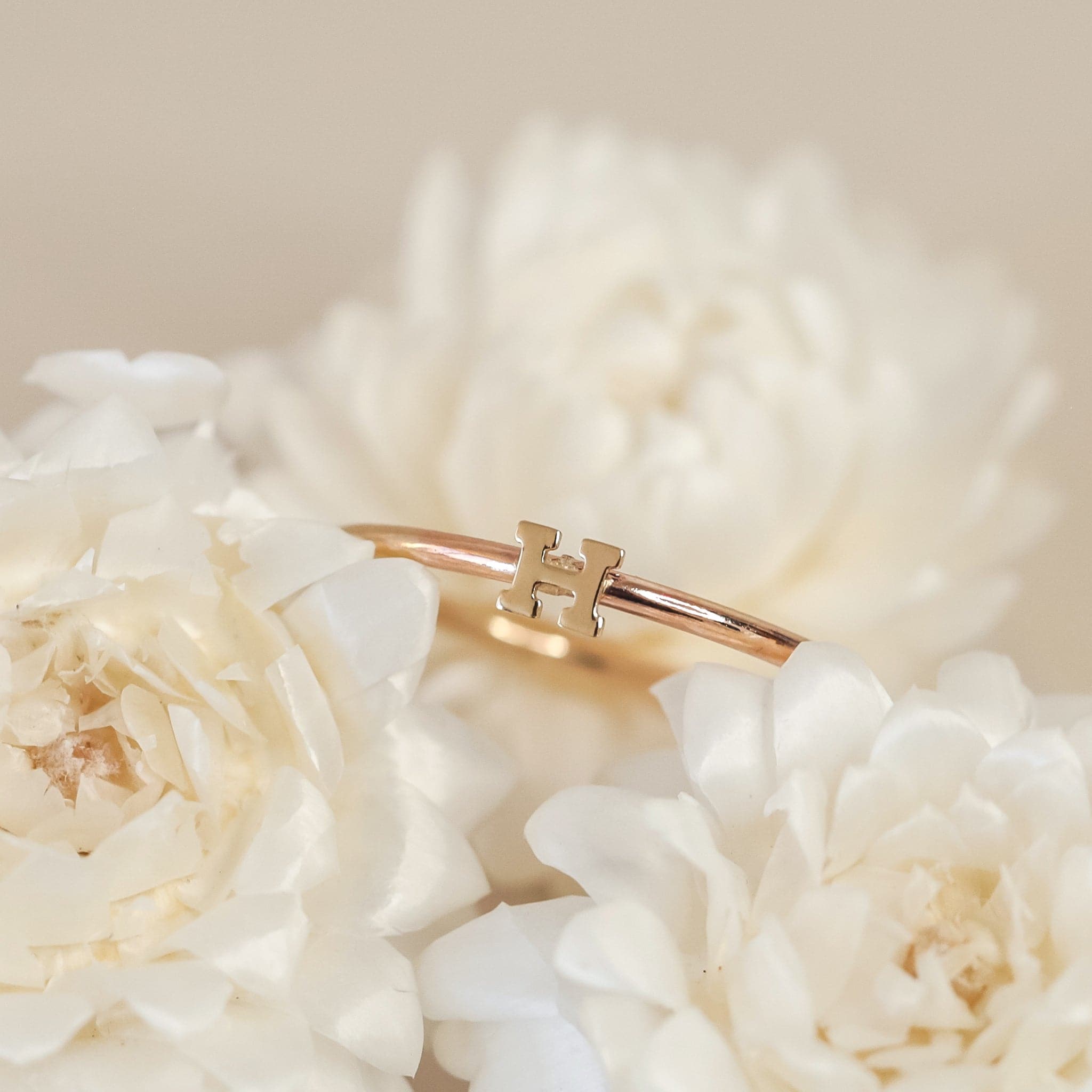 On a cream background is a dainty gold ring with an &quot;H&quot; in the center.
