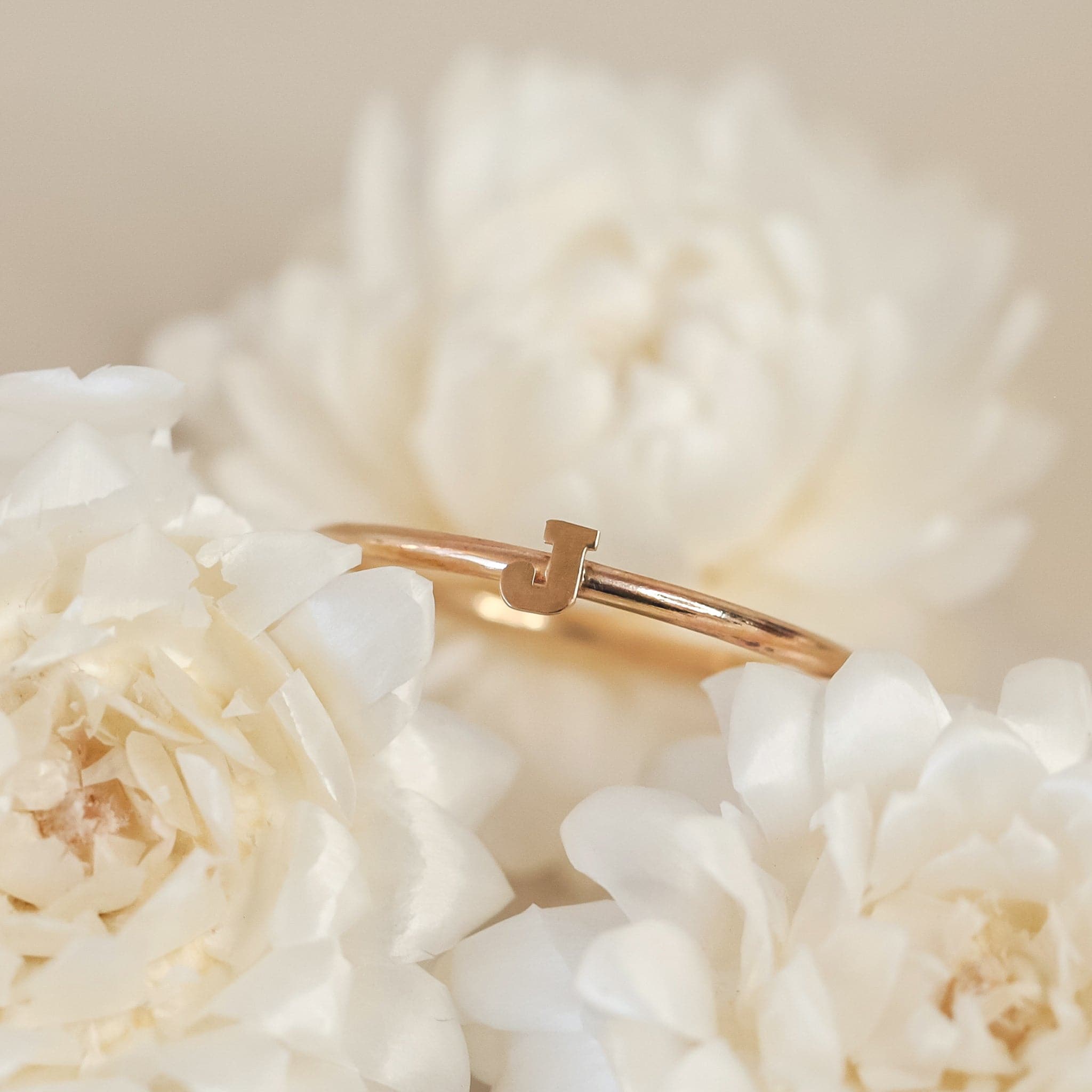 On a cream background is a dainty gold ring with an &quot;J&quot; in the center.