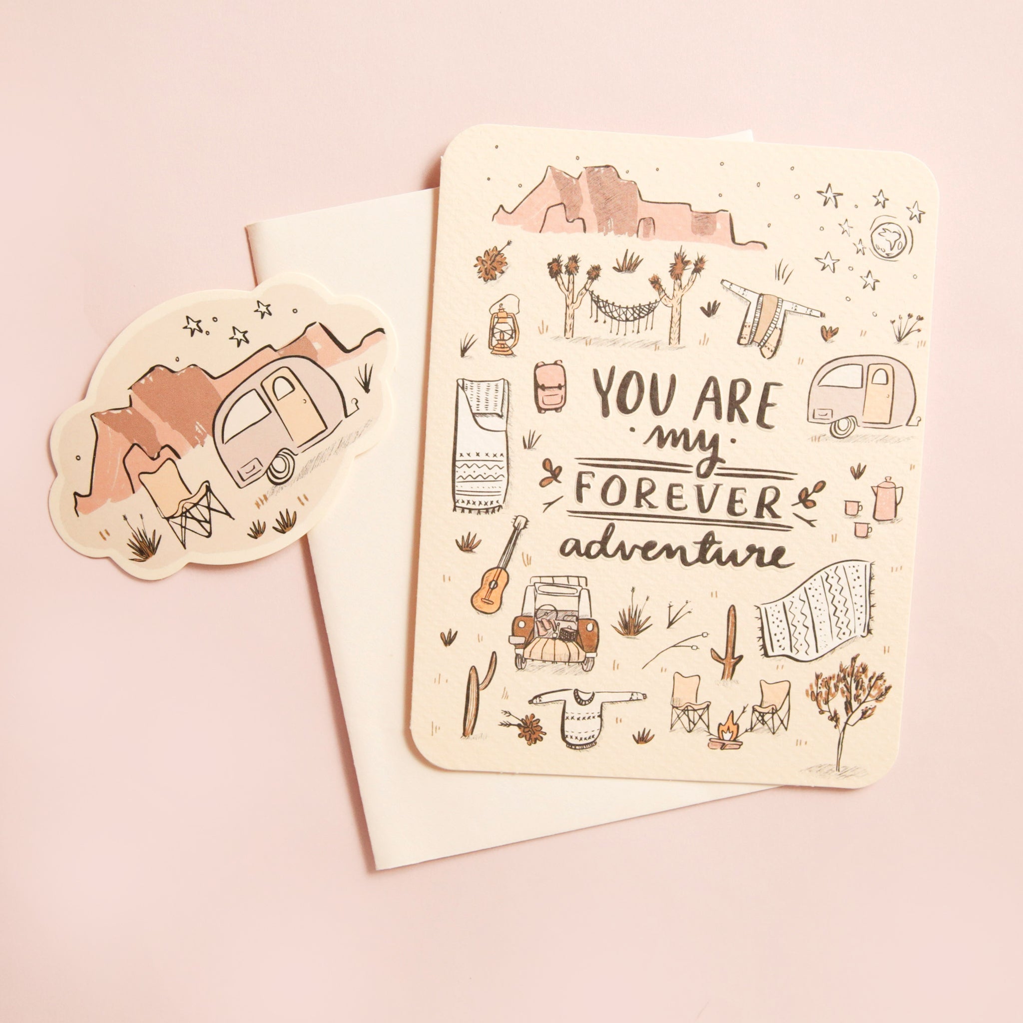 A cream colored card with a bunch of desert illustrations, including Joshua trees, cacti, stars, a camping trailer and more along with the words, "You Are My Forever Adventure". Also included is a white envelope and a small sticker with a camping chair and trailer against a desert mountain range.