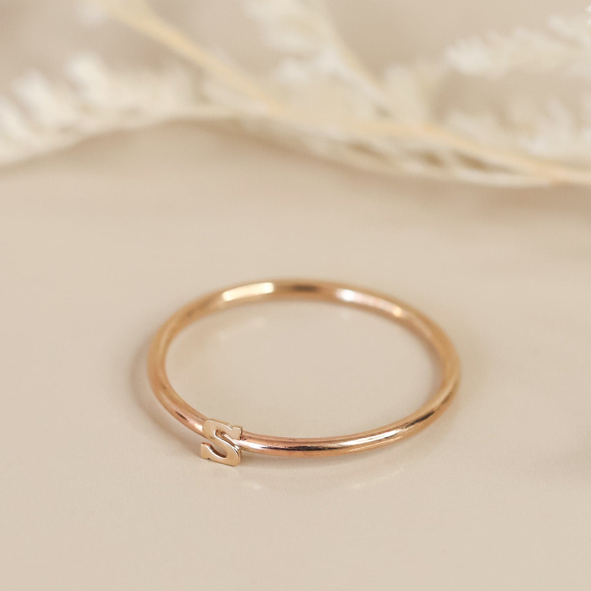 On a cream background is a dainty gold ring with an &quot;S&quot; in the center. 