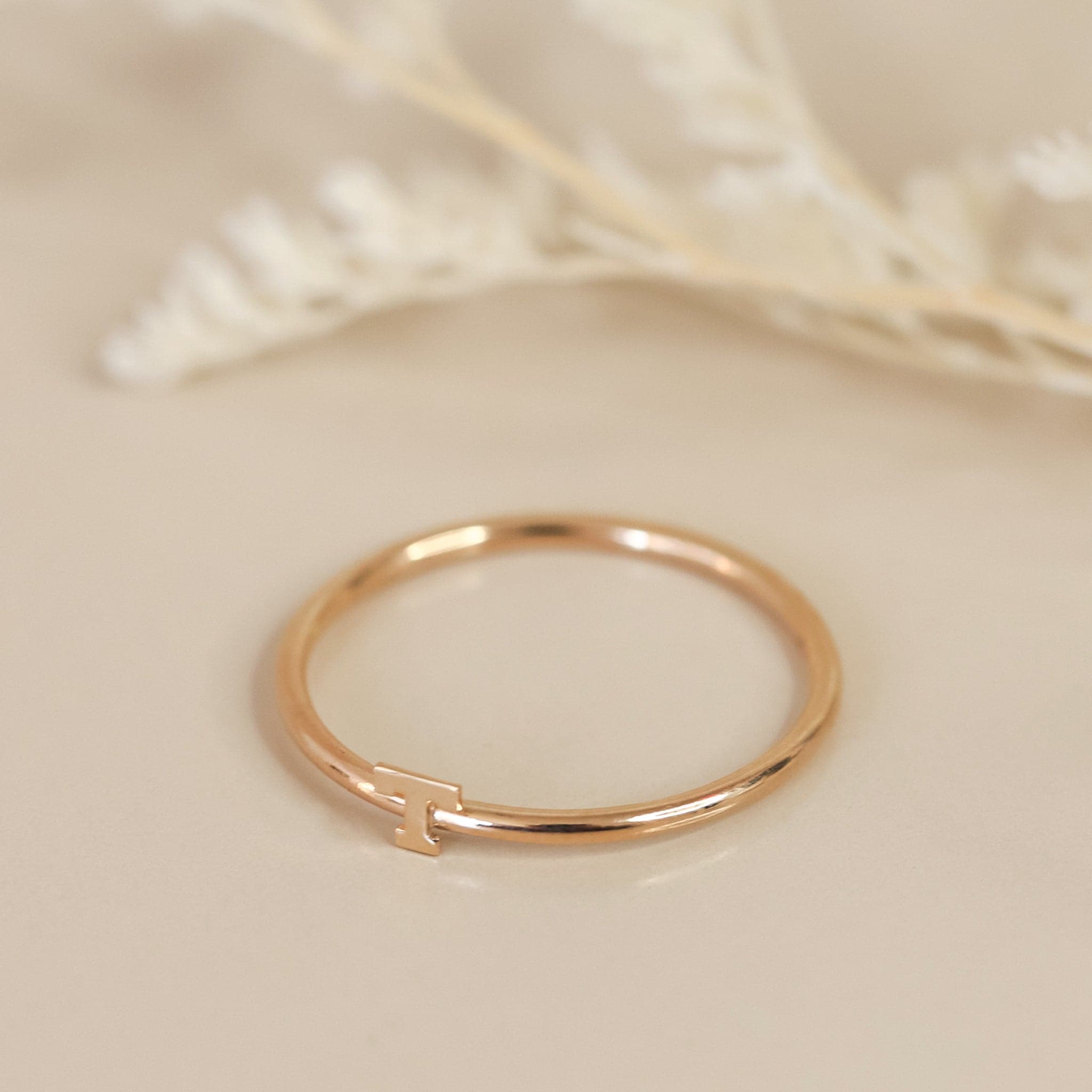 On a cream background is a dainty gold ring with an &quot;T&quot; in the center.