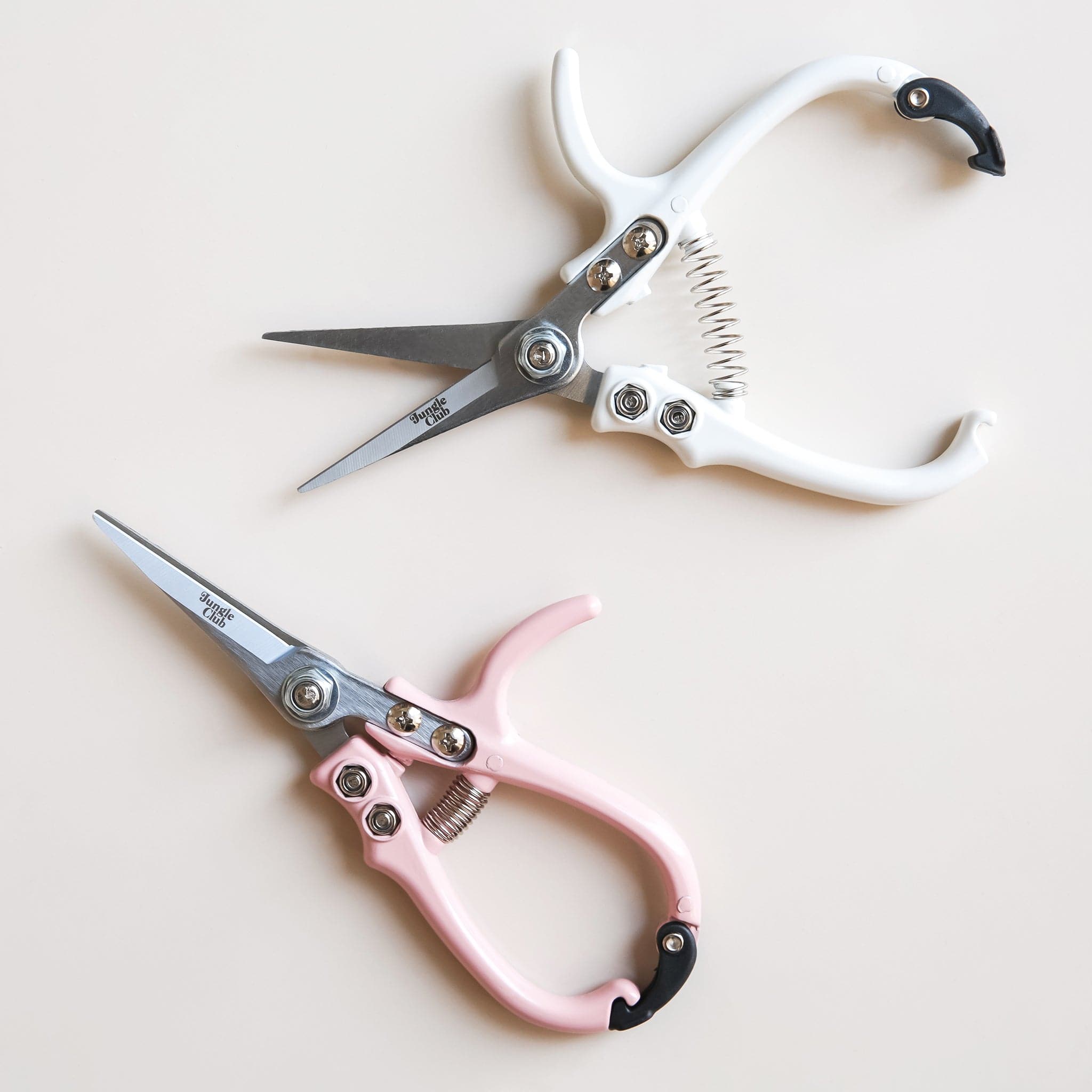 Two pairs of pruning shears laying next to each other. The pair towards the bottom is fasten closed with pastel pink handles and the pair above is positioned unclasped with white handles. 