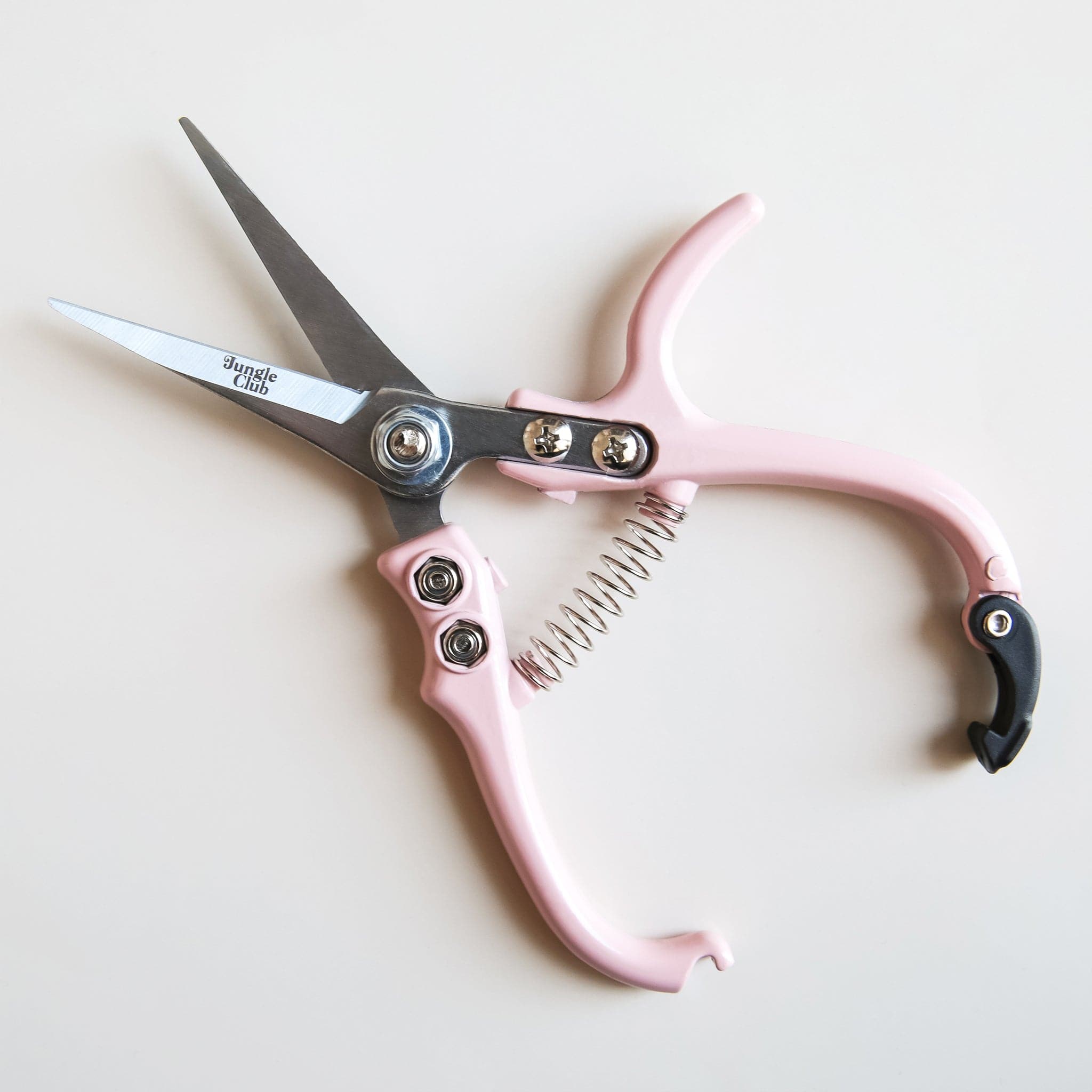 Pair of pruning shears with pastel pink handles and metal blades positioned open. The outer blade reads 'jungle club' in small, playful lettering. The back clasp of the shear handles remain unclipped, allowing for the shear blades to be open. 