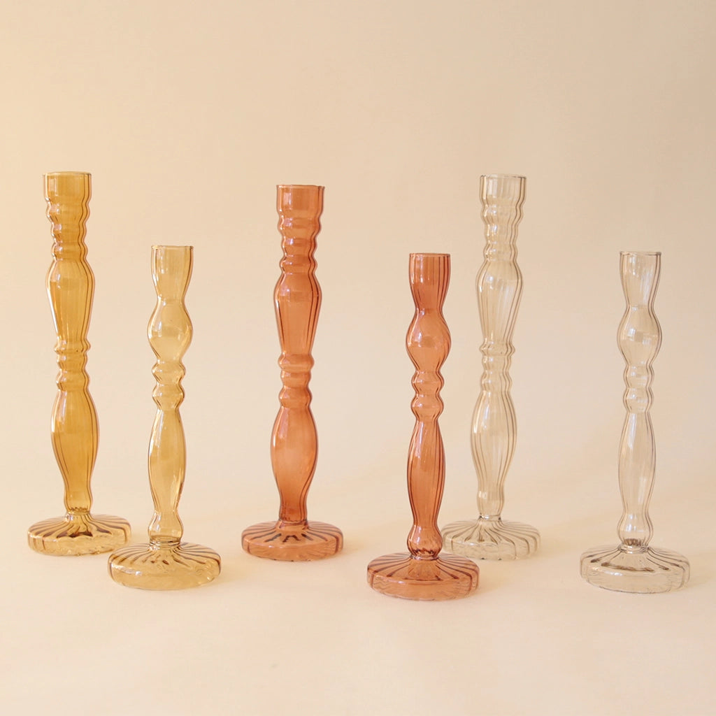 All six bud vases photographed together showing the different sizing and color ways that are available. Each color way has two size options, varying in height and they are available in a mustard yellow, a rust color and a clear smokey color.