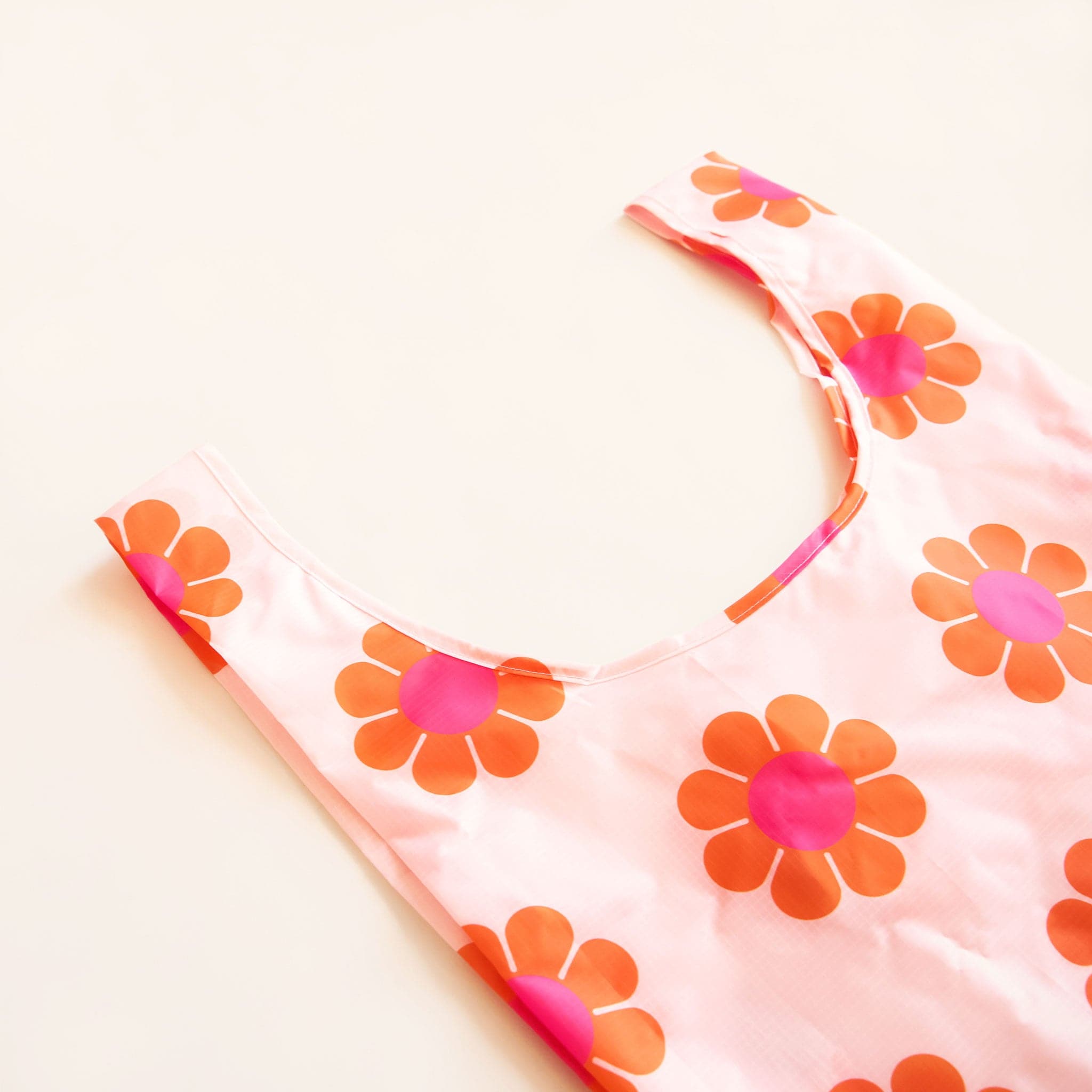 Zoomed in view of peach reusable bag covered in a print of simple flowers with red-orange petals and magenta centers. The bag is positioned flat on a table and has a 'U' shape between two handles.