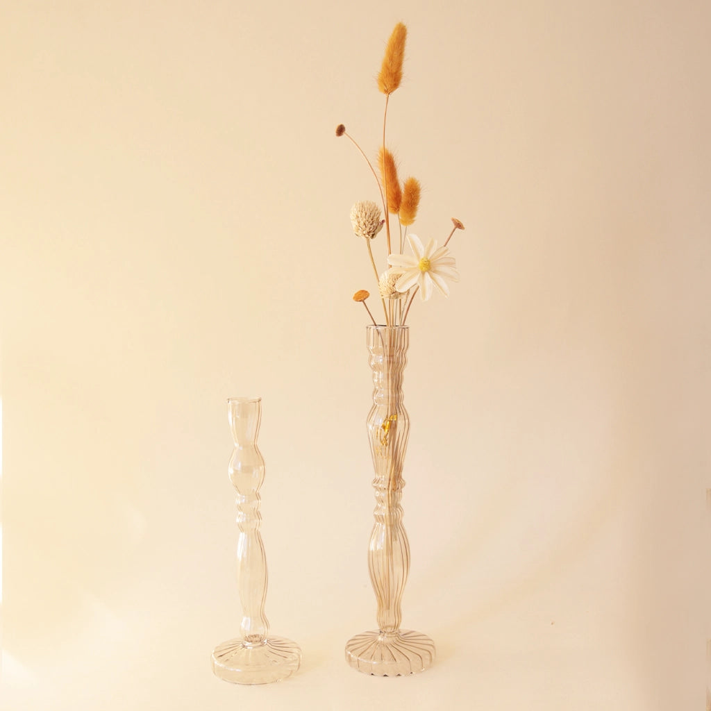 Two slim bud vases with a curvy yet slender design along with vertical ribbing. They are photographed here with a few flower stems placed inside the taller of the two. Each vase sold separately.