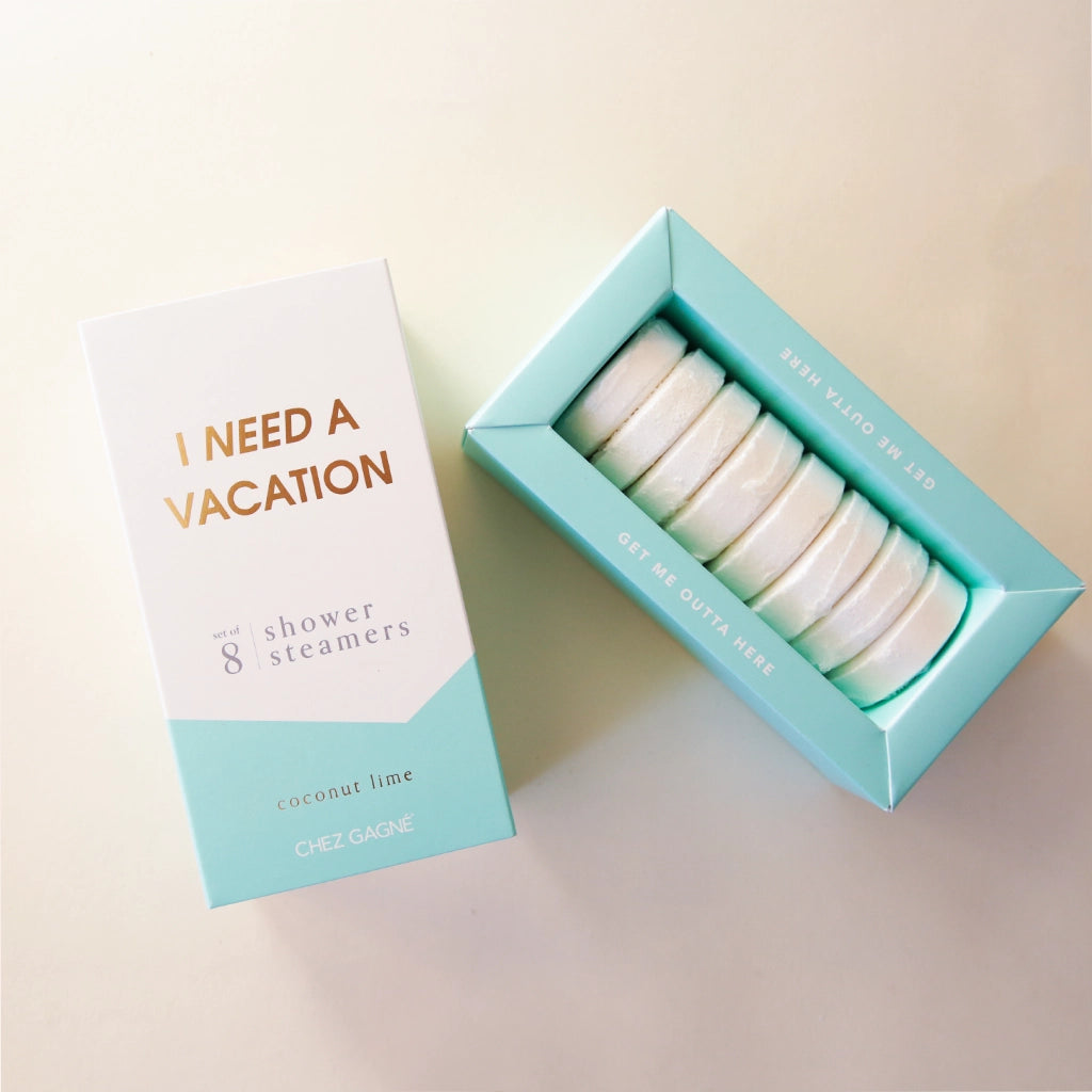 Eight white shower steaming tablets placed inside of a cardboard packaging that is teal blue and white and reads, "I Need A Vacation" in gold text.