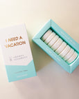 Eight white shower steaming tablets placed inside of a cardboard packaging that is teal blue and white and reads, "I Need A Vacation" in gold text.