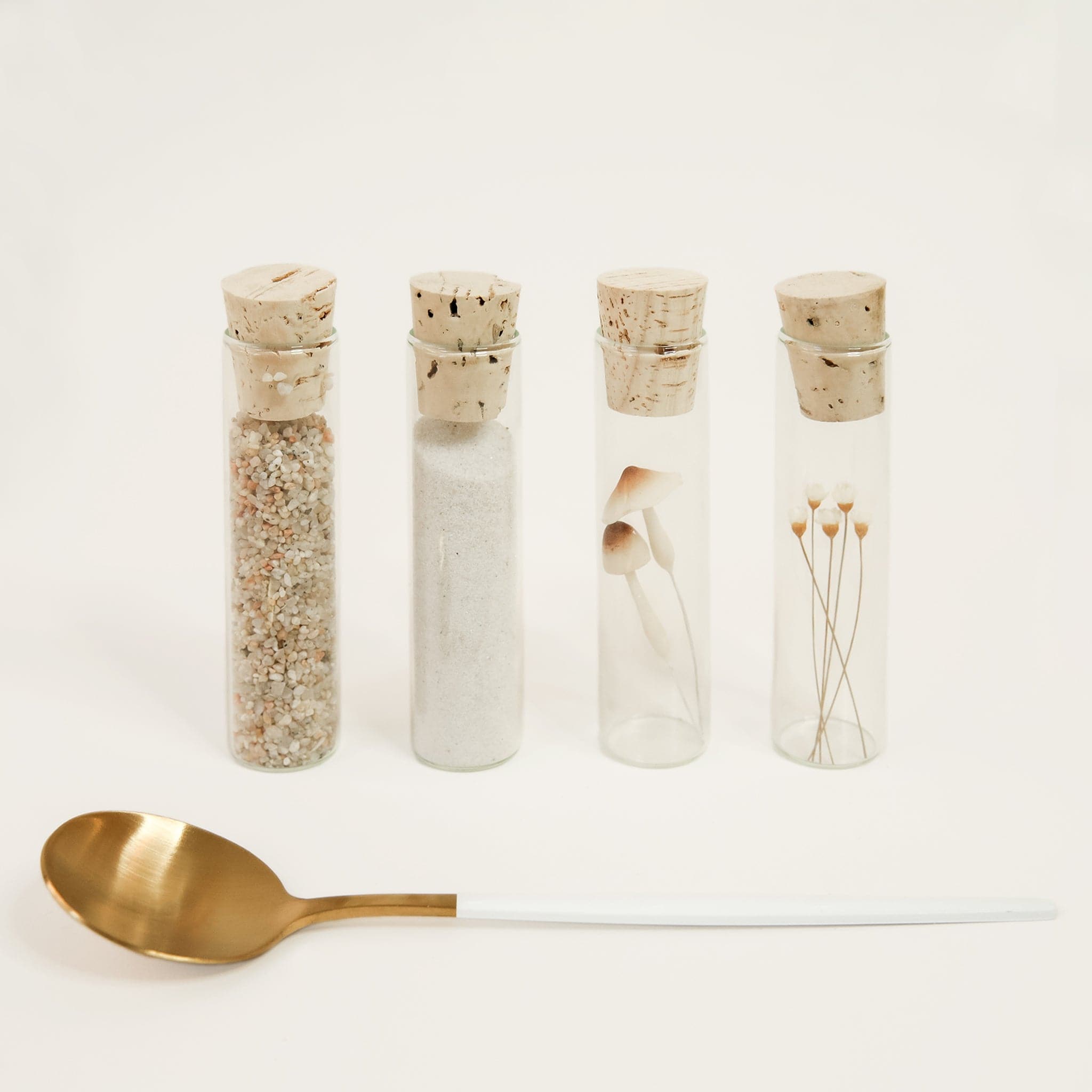 Four glass vials filled with a variety of neutral toned sand, pebbles, mushrooms and dried florals. Each vial is secured closed with light cork. A golden spoon with a long white handle is positioned in front of the vials. 