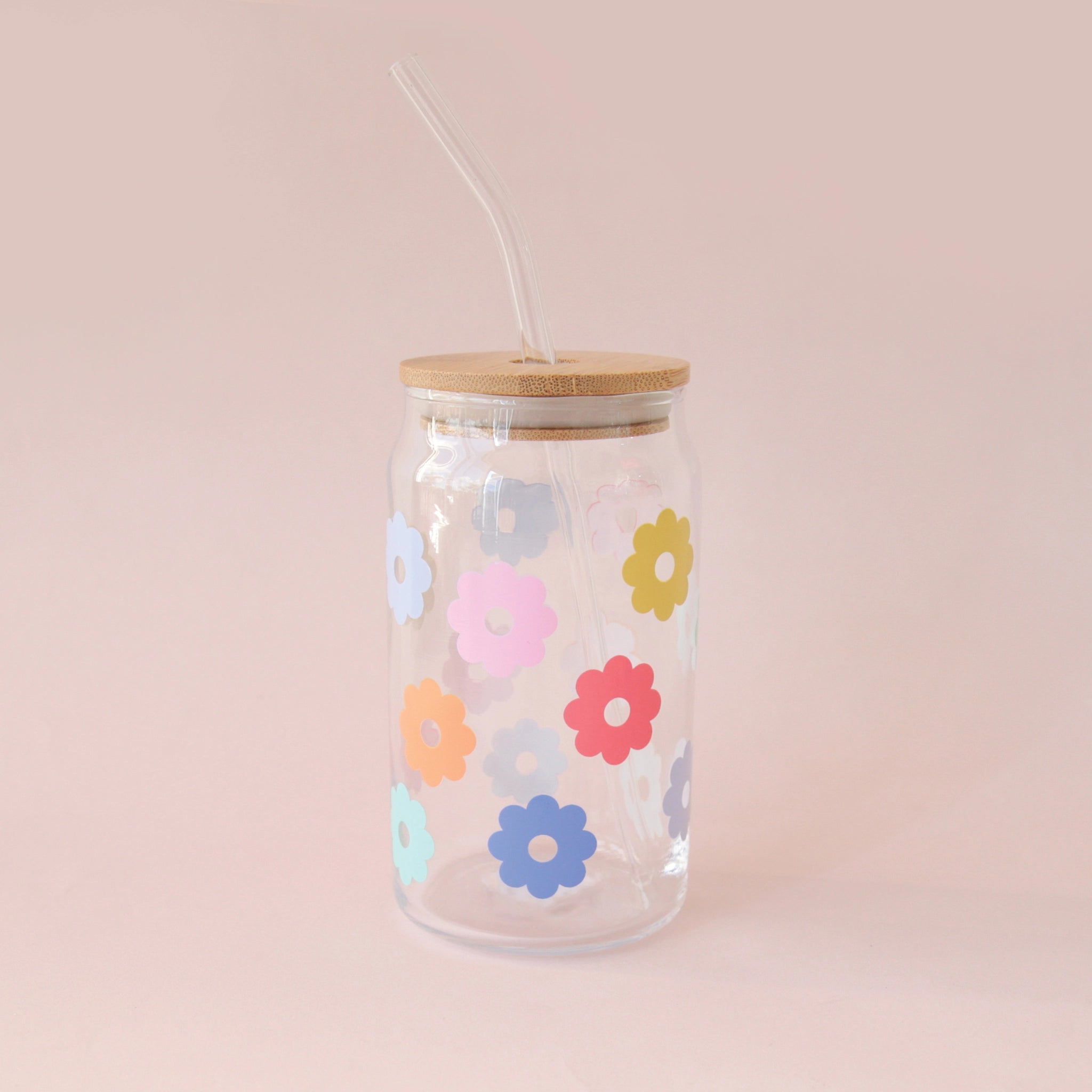 A glass tumbler with multi colored daisies and a wooden lid and a glass straw.