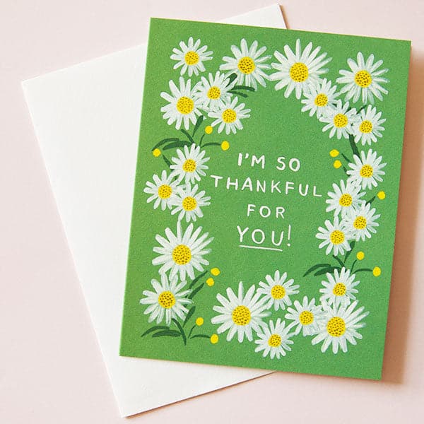 A photograph of a green card and a white envelope behind it. The card has a white daisy border around white text in the center that reads, "I'm So Thankful For You!". 