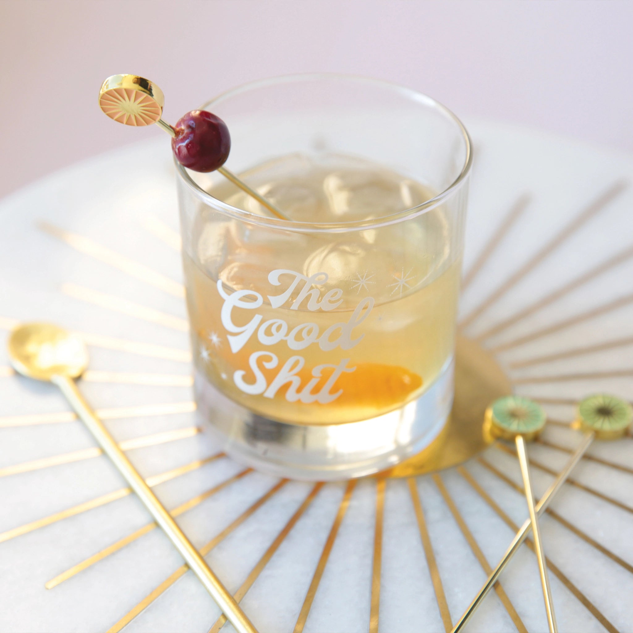 A photograph of a short glass tumbler with a thick bottom and "The Good Shit" printed across the center in white groovy cursive text and staged here with cocktail picks and a cranberry garnish, not included with purchase.