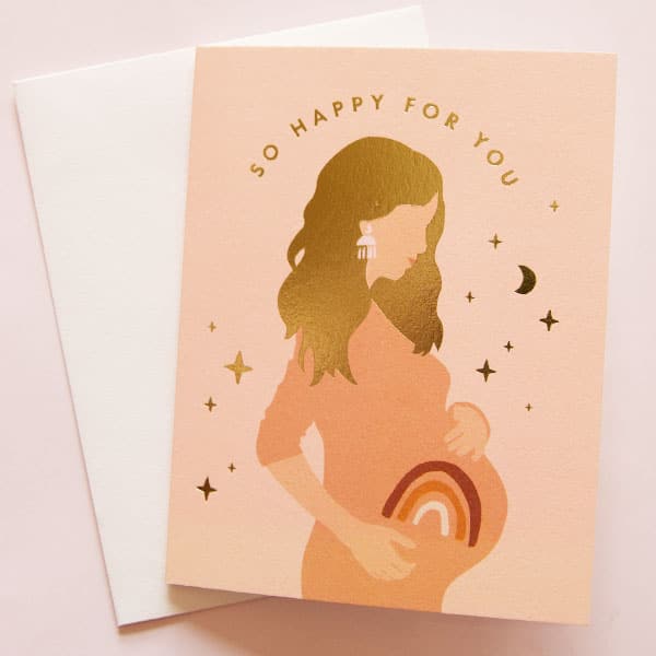 On top of a white envelope is a peach card. In the middle is the drawing of the side profile of a pregnant woman. On her belly is a rust, orange and white rainbow. She has long gold hair. Above her head is gold text that reads ‘so happy for you.’ There are gold stars and moons all around her. 