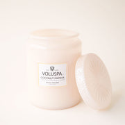 On a cream background is a decorative glass jar candle and a white label that reads, "Voluspa Coconut Papaya" with a glass lid. 
