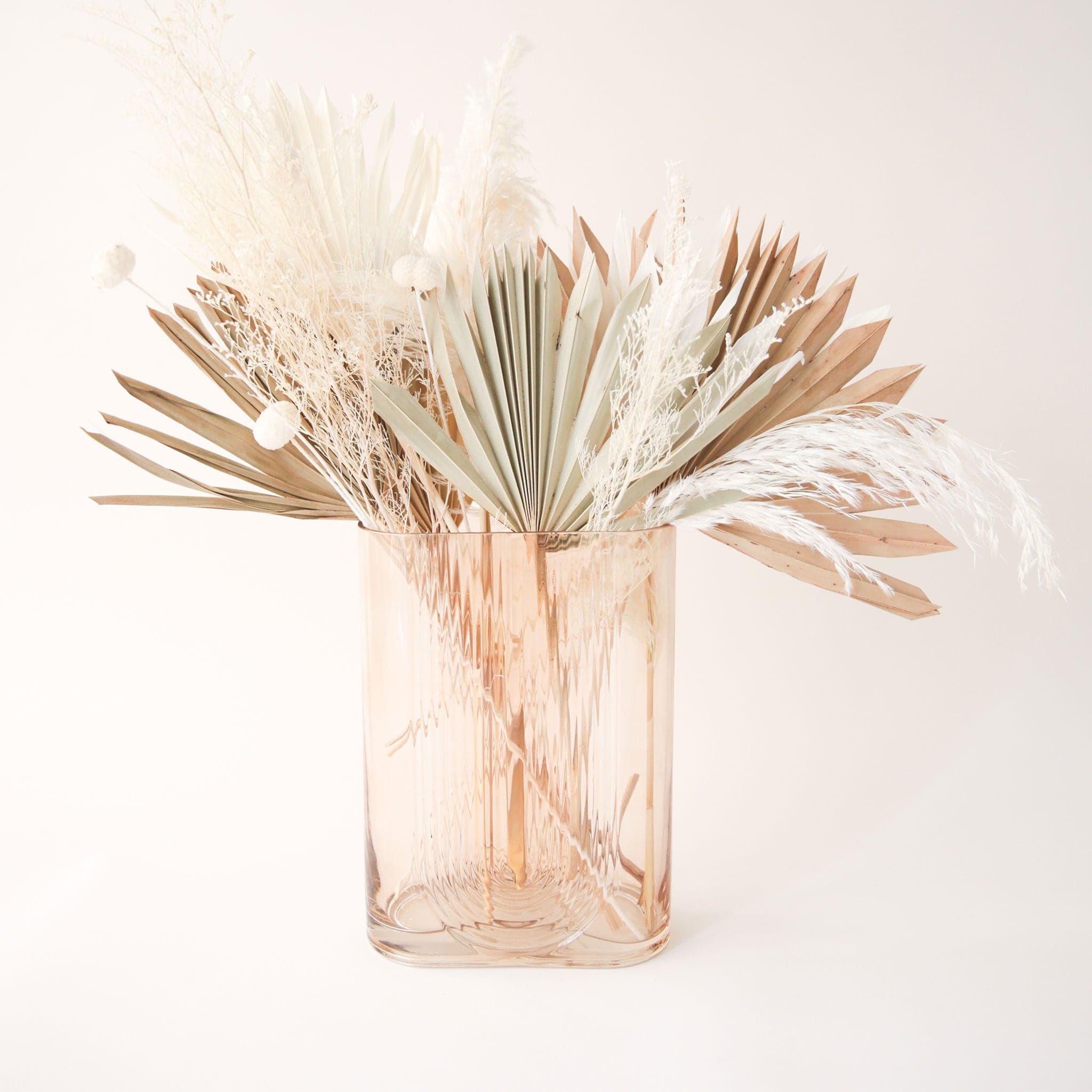 An oval shaped glass vase in a tan shade and an upside down rainbow etching on the front and back filled with dried florals and palm leaves.
