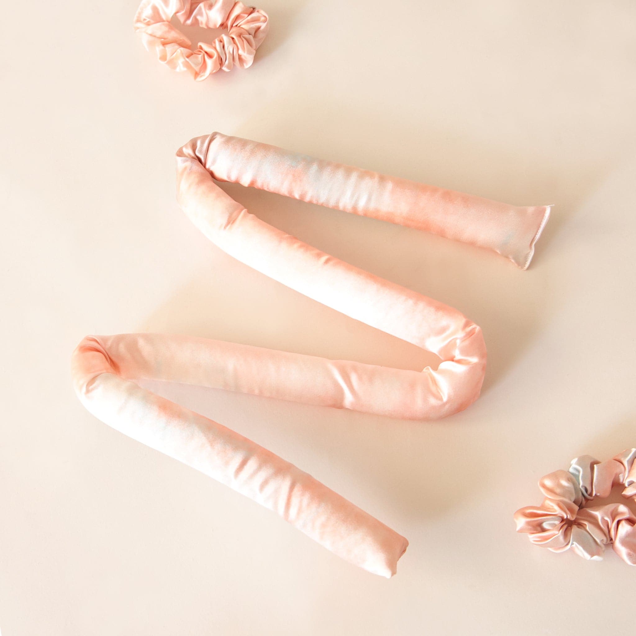Birds eye view of a white background with a blush pink satin covered foam tube. The piece of foam is laying in the middle of the picture and is manipulated in a zig zag pattern. Above and below it is two bush pink satin scrunchies. 