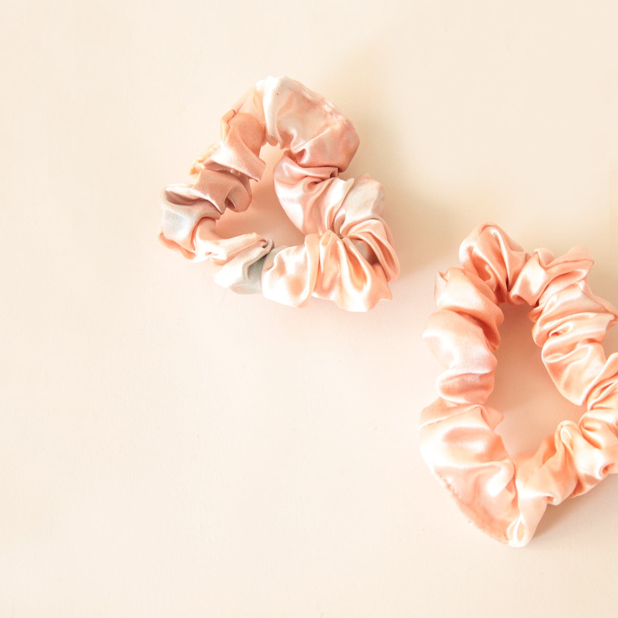 In front of a soft pink background is two blush pink satin scrunchies.