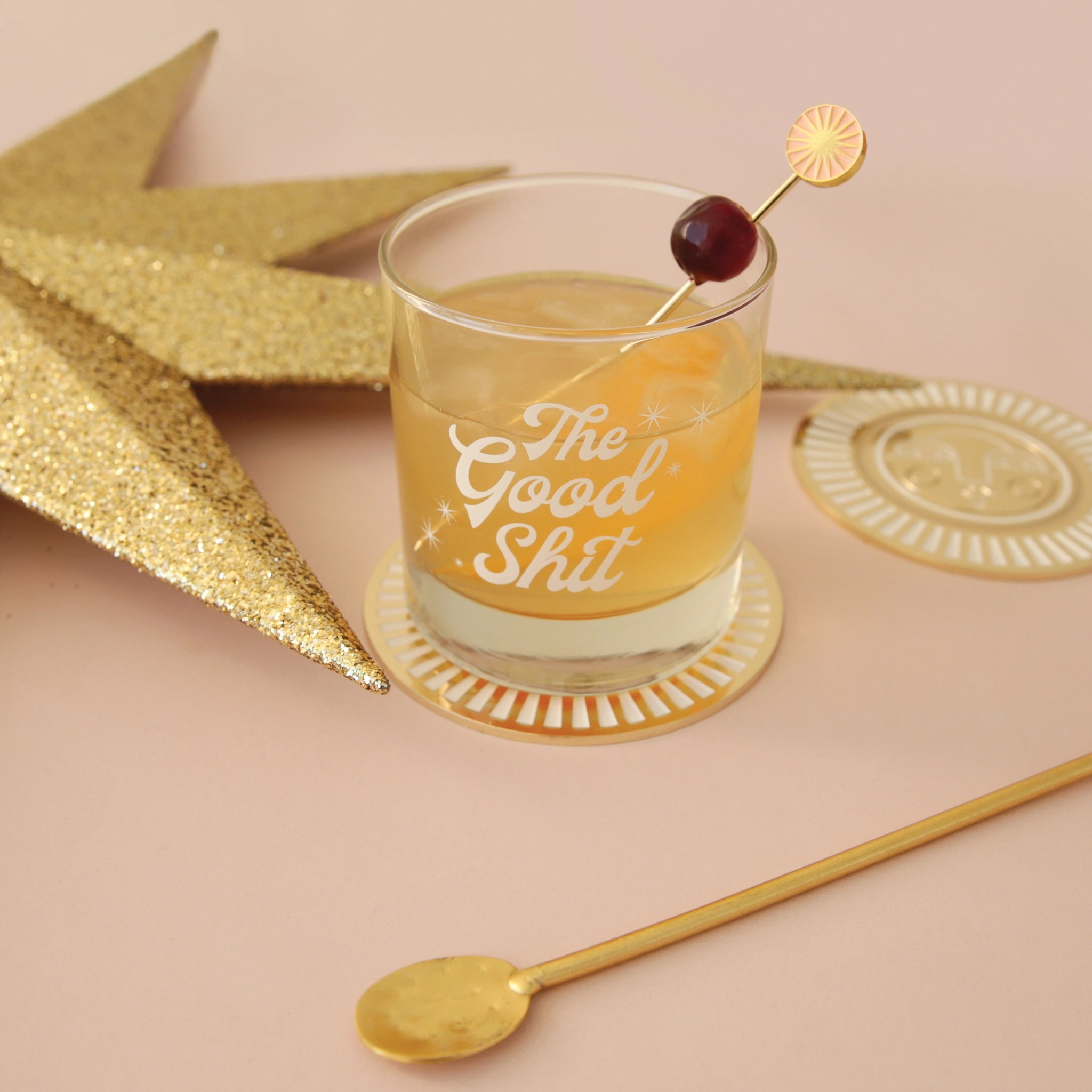 A photograph of a short glass tumbler with a thick bottom and &quot;The Good Shit&quot; printed across the center in white groovy cursive text and staged here with cocktail picks and a cranberry garnish, not included with purchase.\