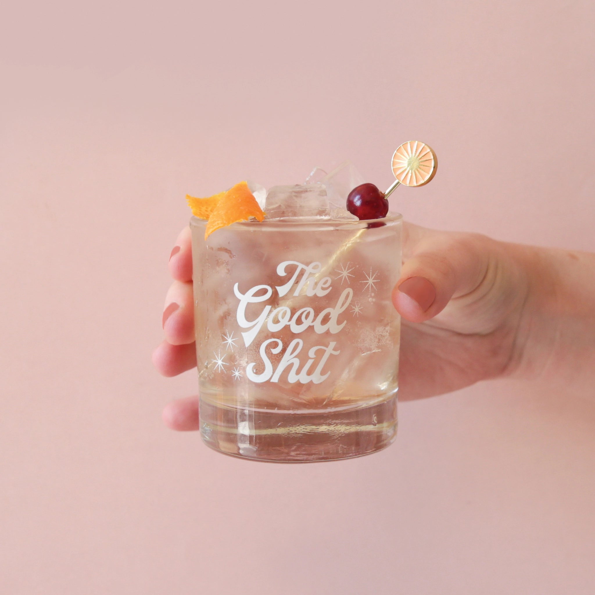 Against a peachy pink background is a photograph of a short glass tumbler with a thick bottom and &quot;The Good Shit&quot; printed across the center in white groovy cursive text staged here with a cocktail pick along with a cranberry and orange peel garnish.