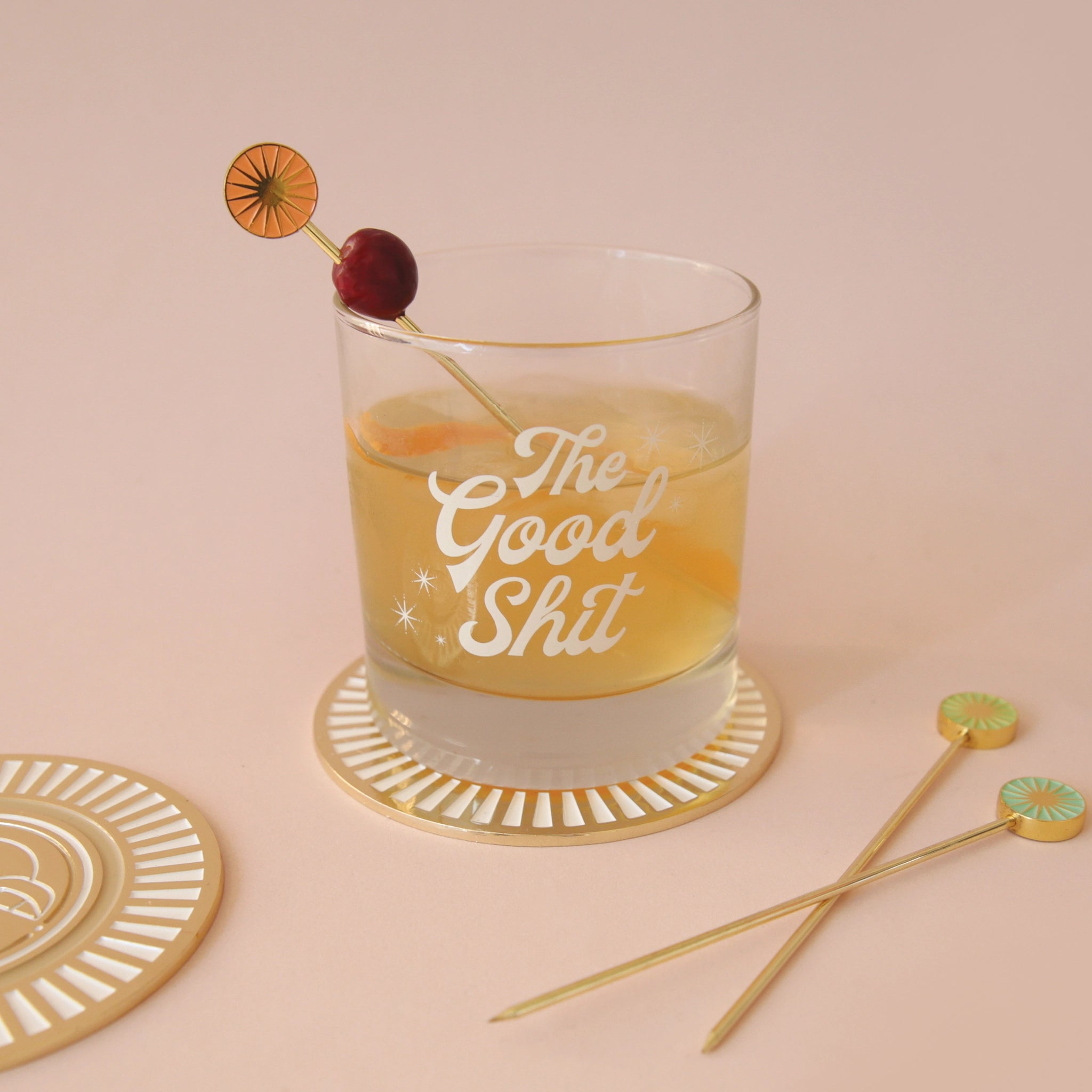 A photograph of a short glass tumbler with a thick bottom and &quot;The Good Shit&quot; printed across the center in white groovy cursive text and staged here with cocktail picks and a cranberry garnish, not included with purchase.